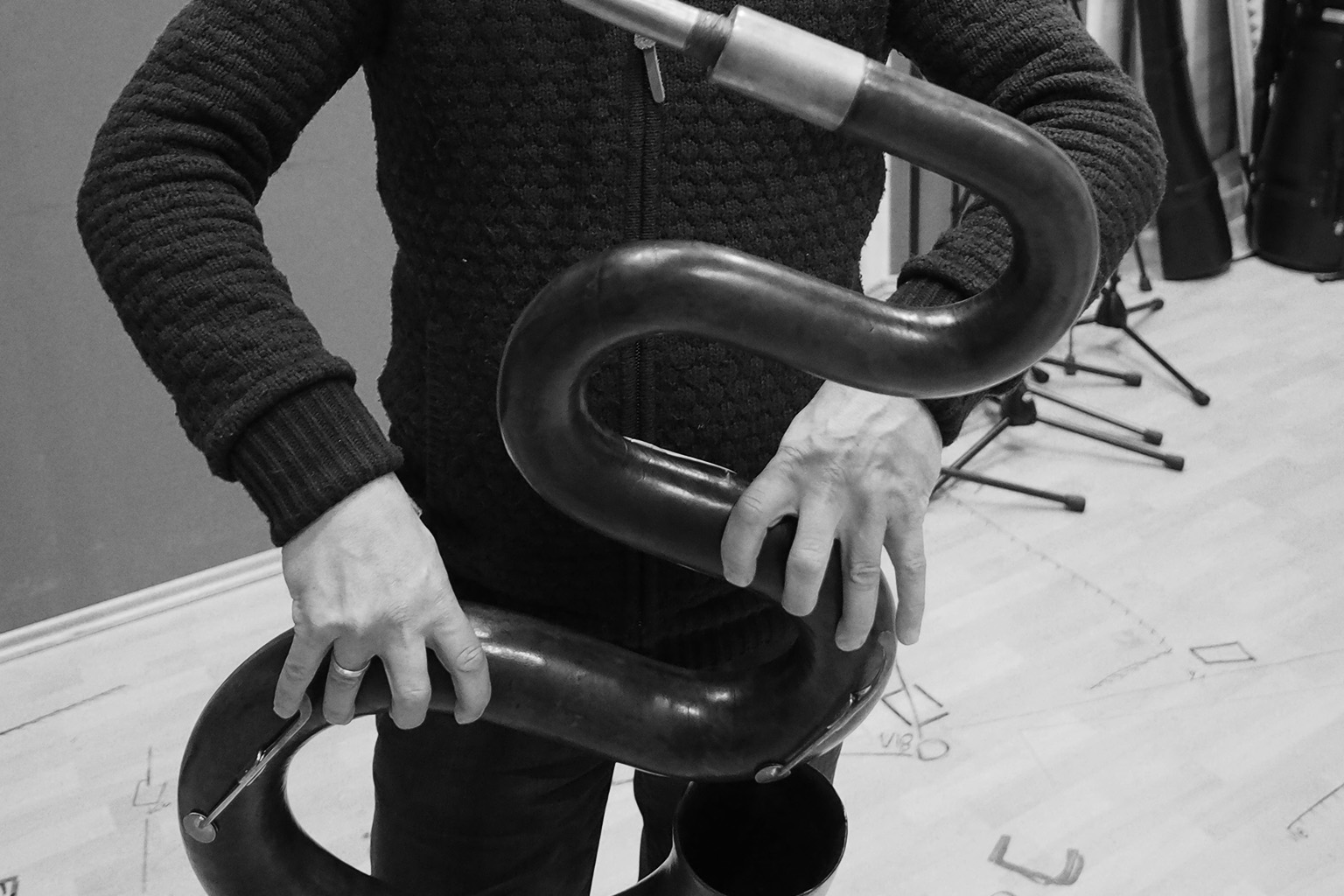 Black and white image of a person holding a large, dark, S-shaped musical instrument, likely an oversized wind instrument or a unique horn, with hands positioned on its curved body.