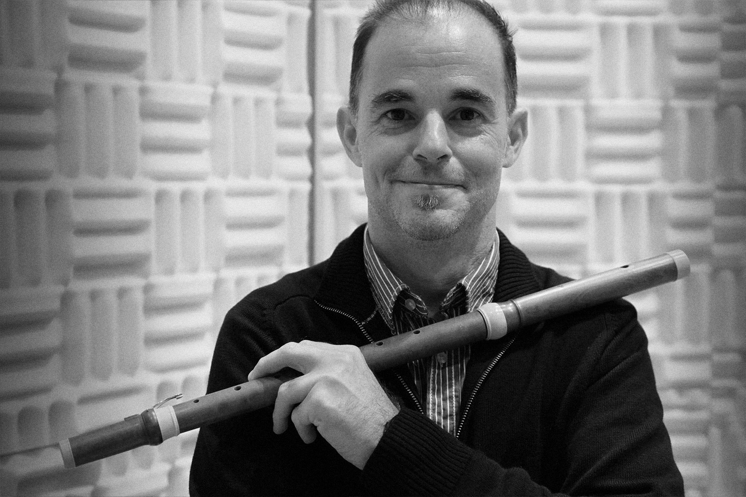 Black and white portrait of a smiling man holding a wooden flute in a music studio