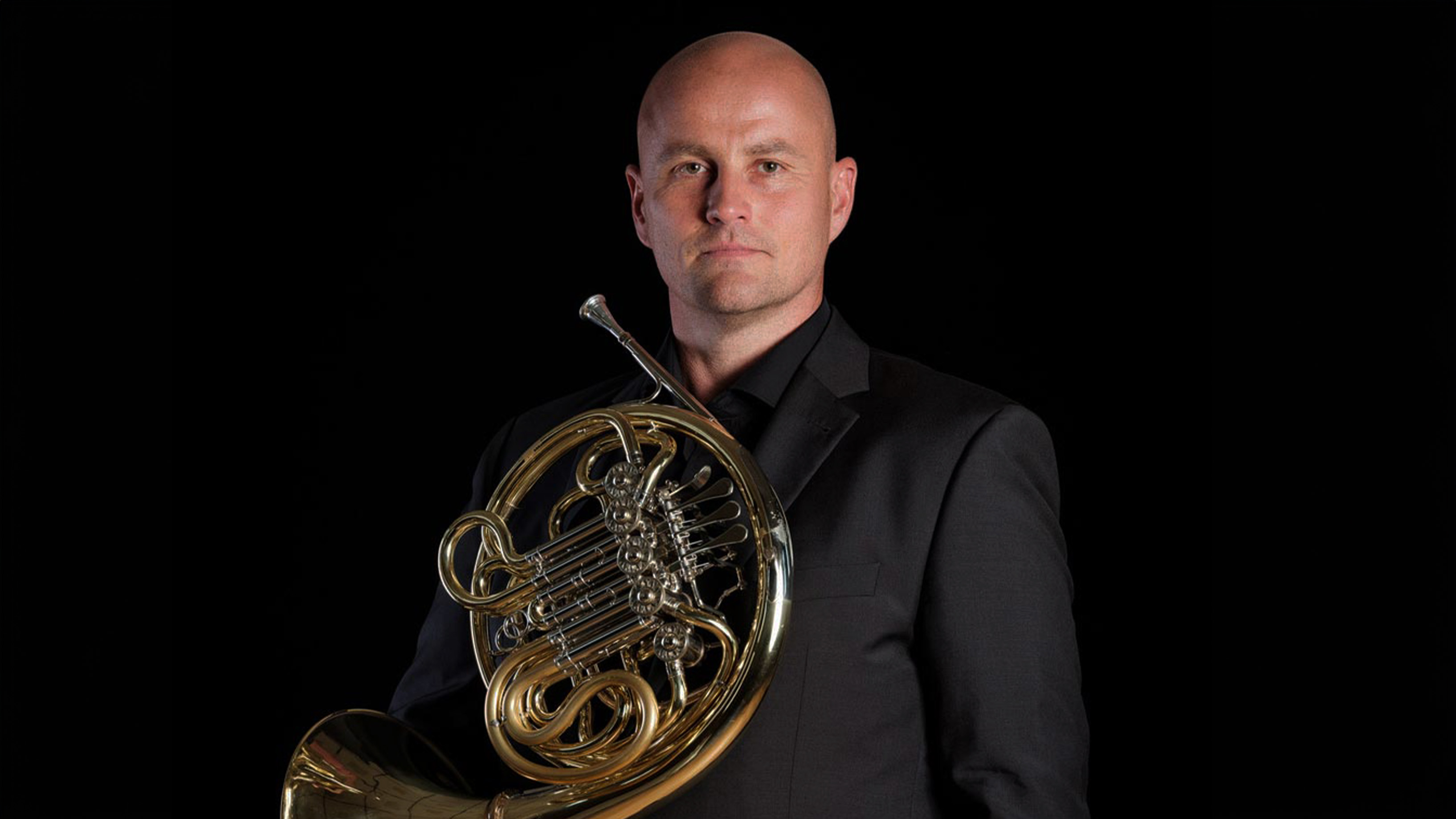 Bald man in a black suit holding a French horn.