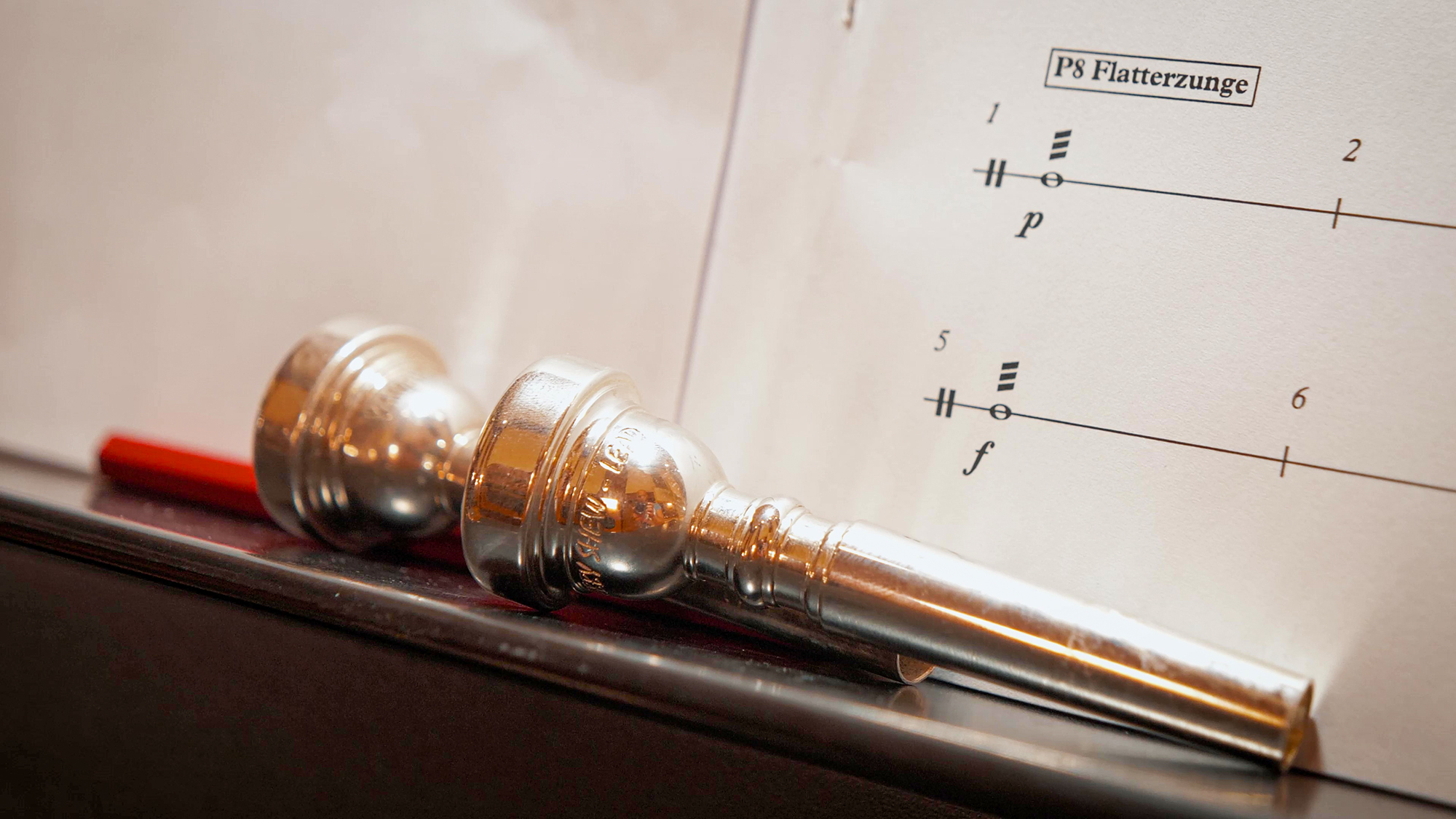 Two silver trumpet mouthpieces and a red pencil lie on a black surface, with a sheet of sheet music featuring ''P8 Flatterzunge'' in the background.