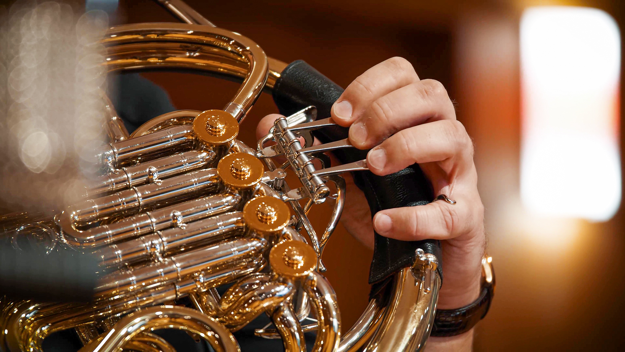Musician's hand pressing keys on a French horn, showcasing the instrument's brass and silver details during a performance.