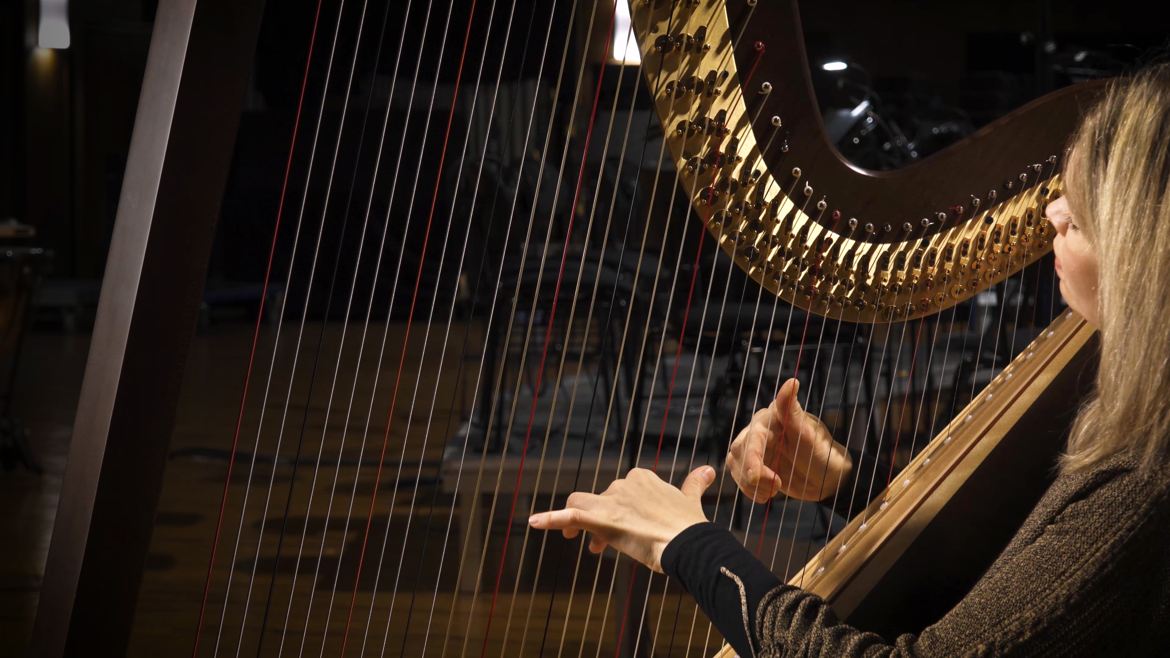 Woman playing a golden pedal harp in a darkened room, fingers on the strings.