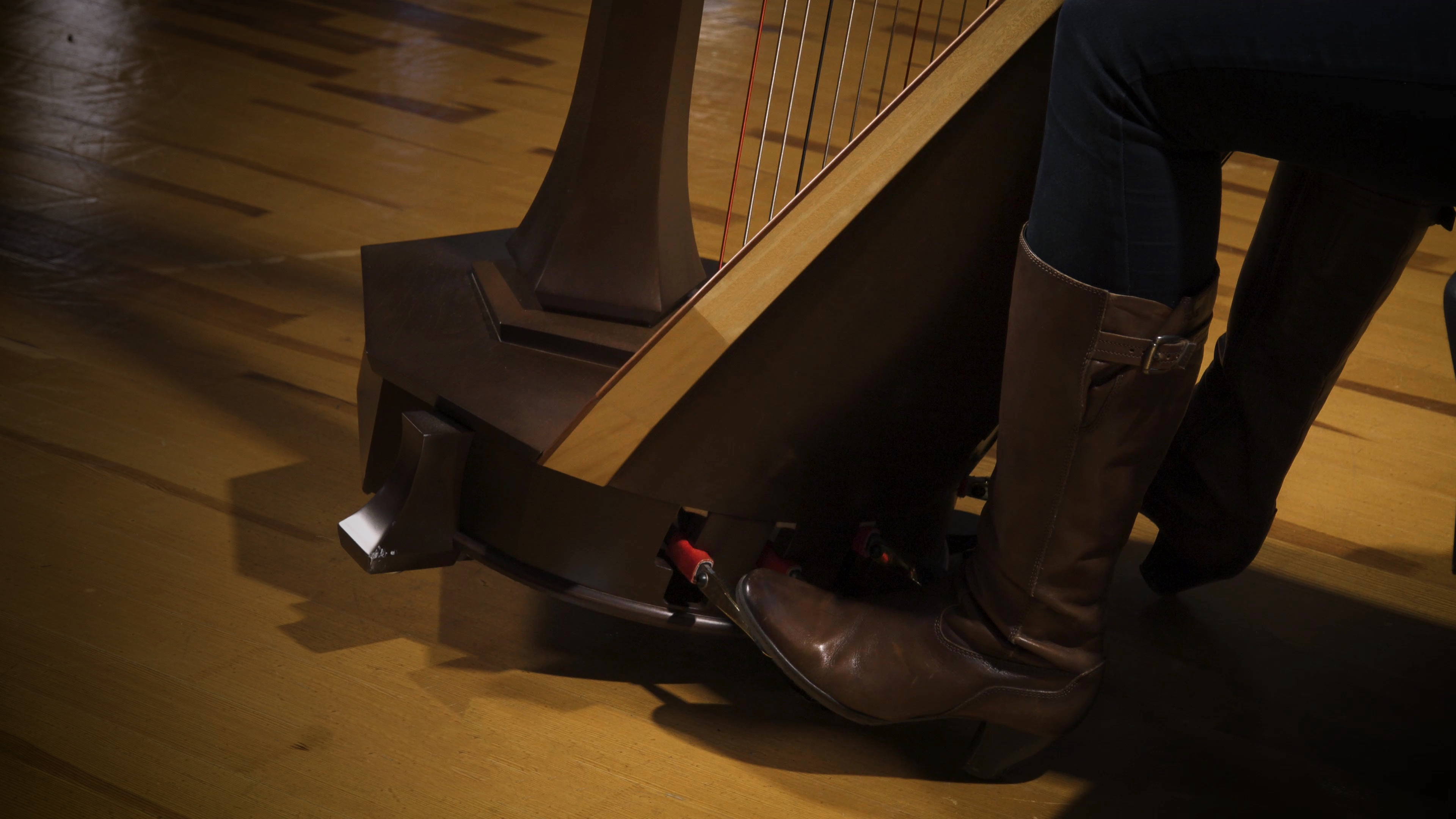 Musician's foot in a brown leather boot operating pedals on a harp, set on a polished wooden floor.