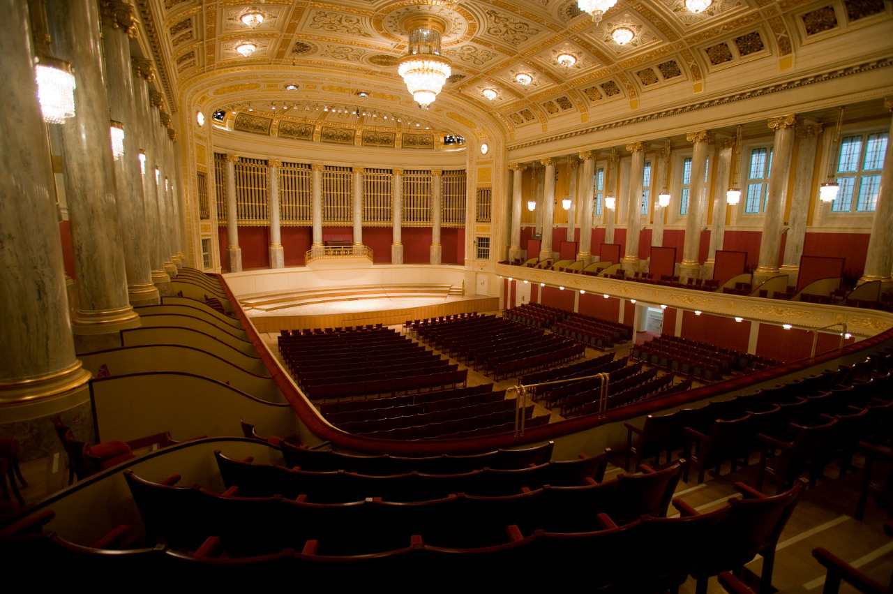 Grand empty concert hall or opera house with ornate gold and white ceiling, marble columns, red walls, wooden stage, and rows of dark red velvet seats viewed from an upper balcony.
