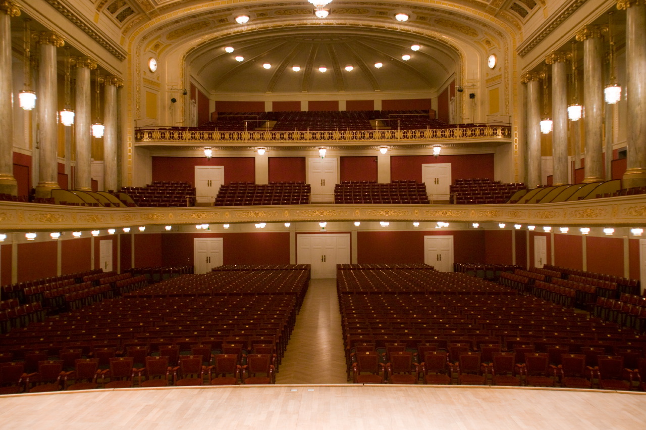 Grand, empty concert hall or theater interior with rows of plush red seats, ornate gold balcony railings, elegant white columns, and illuminated dome ceiling.