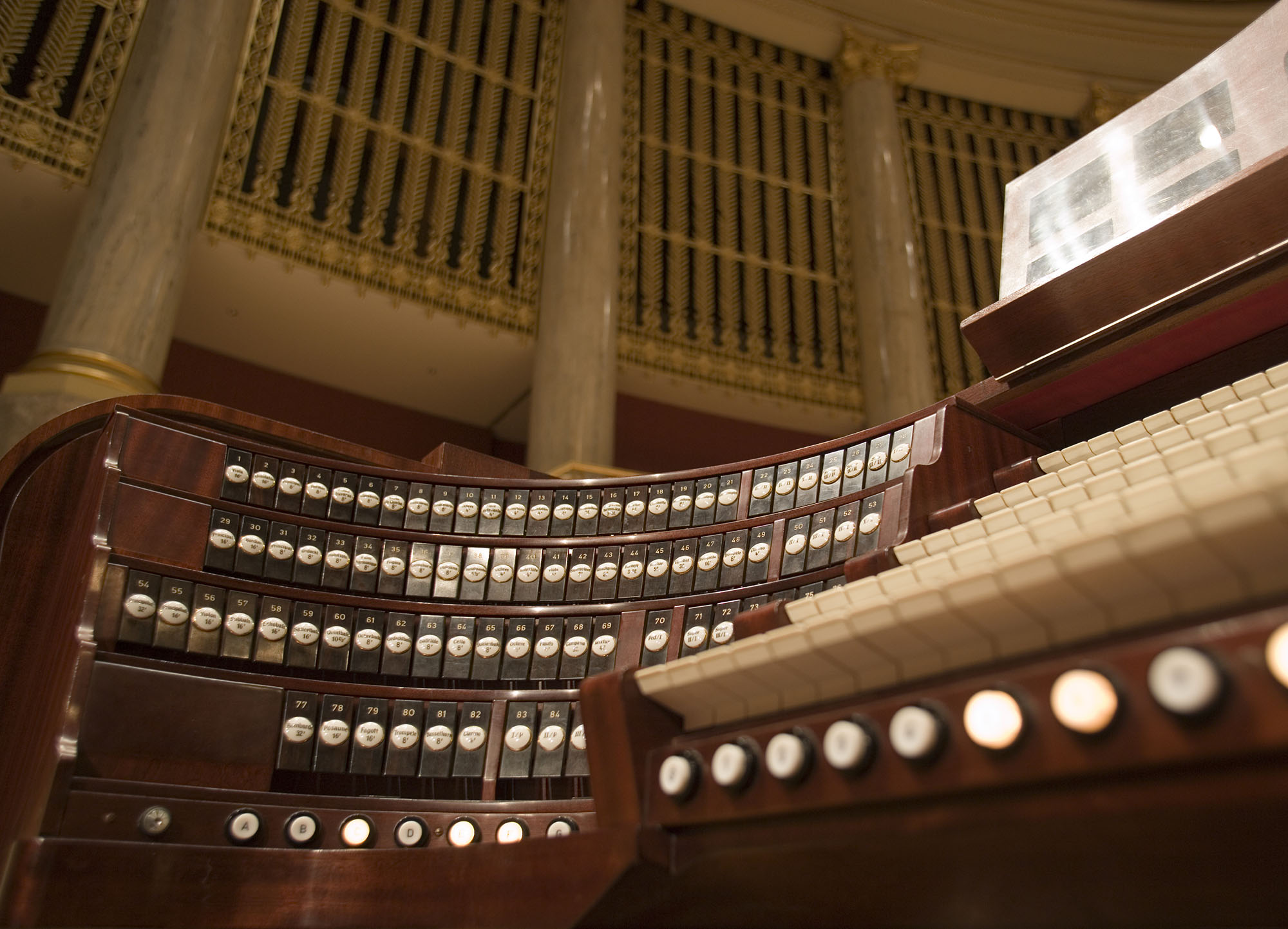 Close-up of a grand pipe organ's wooden console, featuring multiple rows of numbered and labeled stop keys, white keys, and decorative gold and marble structural elements in the background, highlighting the craftsmanship of musical instruments and architecture.
