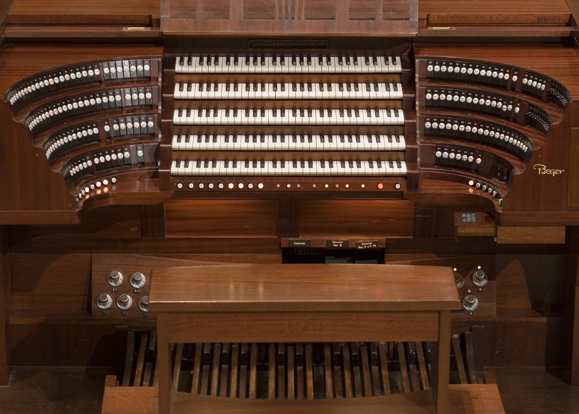 Full view of a large, brown wooden Rieger pipe organ console, showing multiple rows of white and black keys, numerous stop tabs on curved stacked panels, foot pedals, and expression pedals.