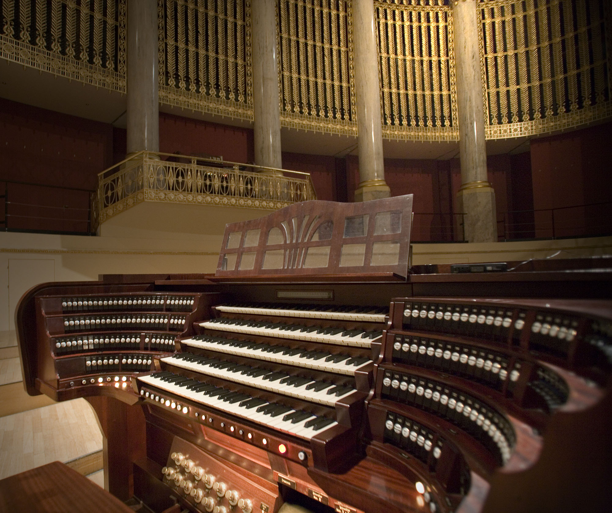 Grand pipe organ console with multiple keyboards and stops, dark wood finish, golden ornate balcony railings and pillars in a dimly lit auditorium.