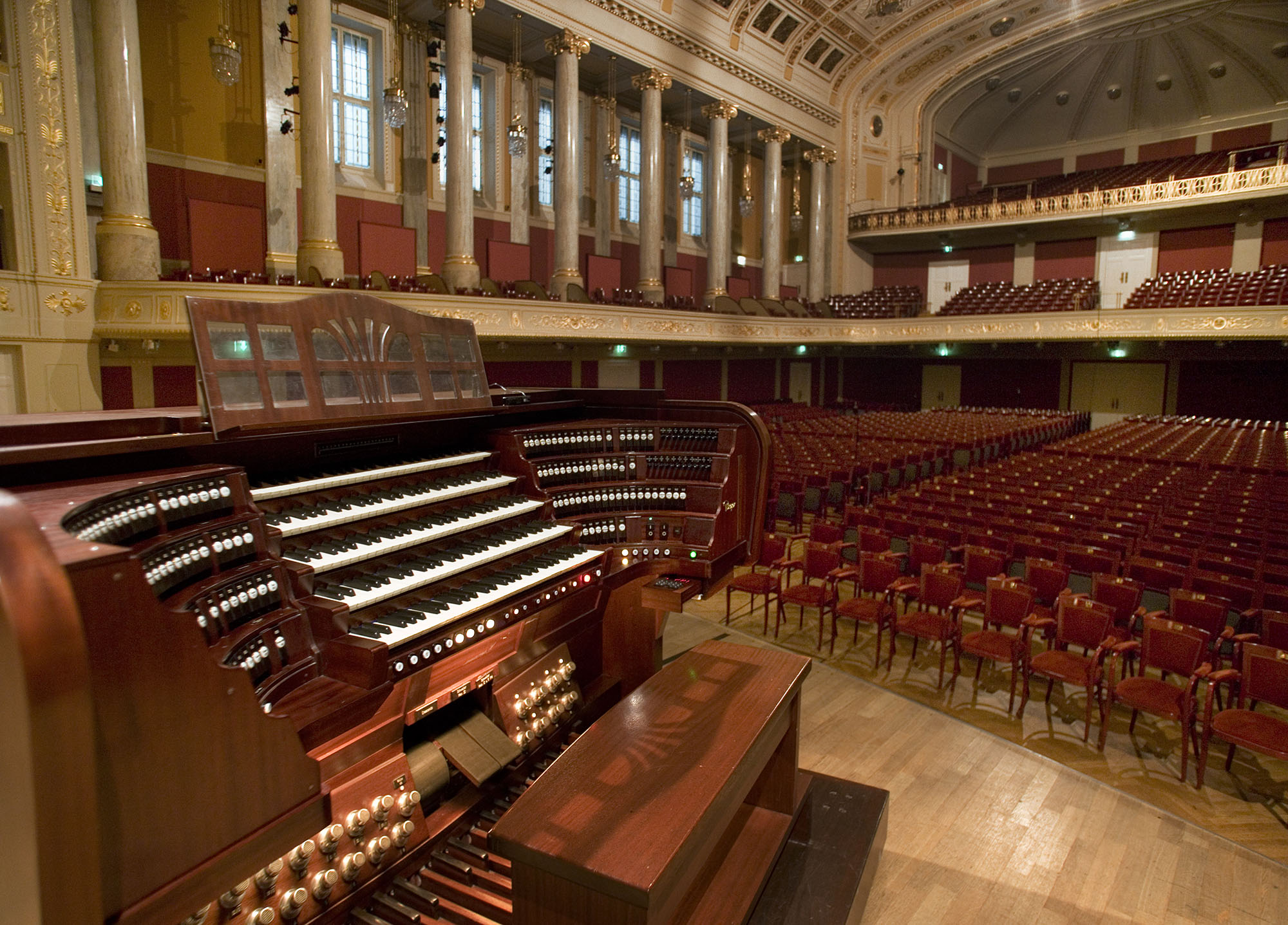 Grand pipe organ console with multiple keyboards and stops, positioned on a wooden stage within a large, elegant concert hall or auditorium filled with rows of empty red seats.