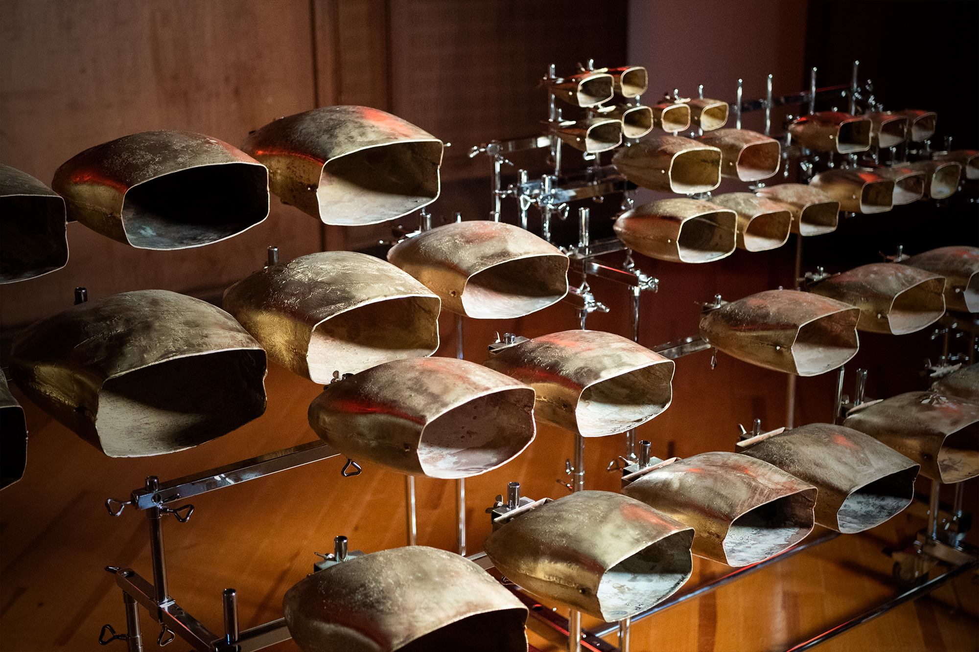 Multiple rows of large, bronze-colored cowbells or agogo bells mounted on metal stands, creating a musical instrument display