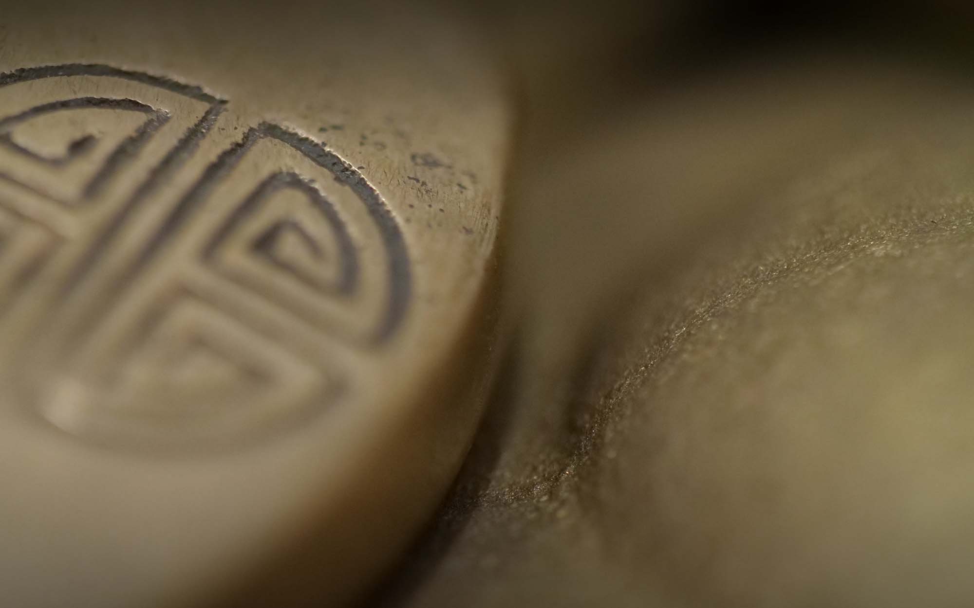 Close-up focused image of an engraved metal surface with an intricate pattern on the left, next to a blurry, textured, golden-brown surface on the right.