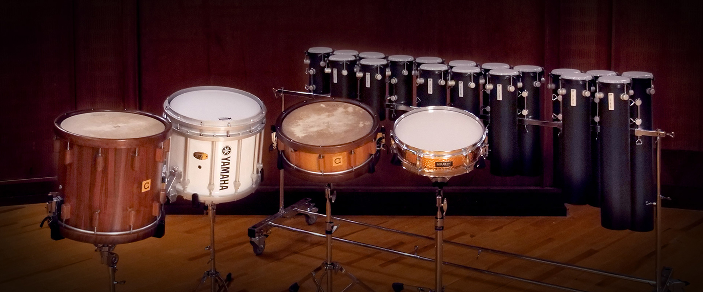A collection of percussion instruments, including a large wooden drum, a white Yamaha snare drum, a copper Kolberg snare drum, and multiple tall, black tube drums with grey tops, all set up on stands on a wooden floor in a studio or concert hall.