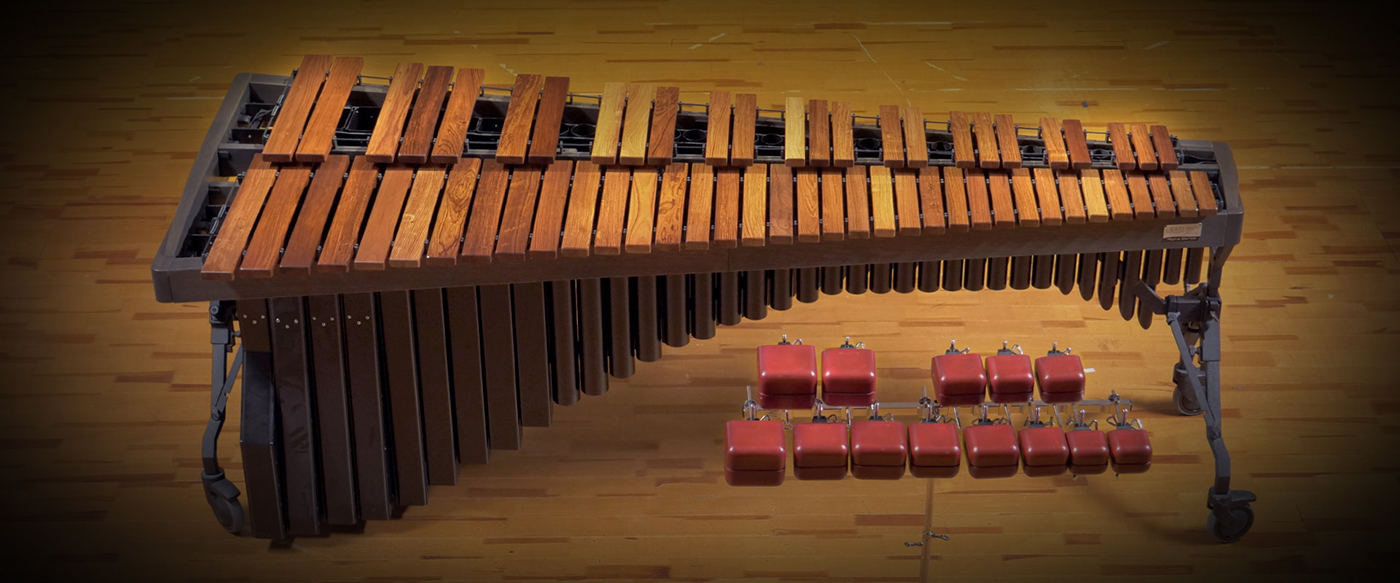 Full overhead view of a large, professional-grade marimba with light brown wooden bars, dark brown resonators, and a dark grey stand. Below the keyboard, a rack of nine red square-shaped blocks with white attachment clips are visible. The marimba rests on a light brown wooden floor with visible grain.