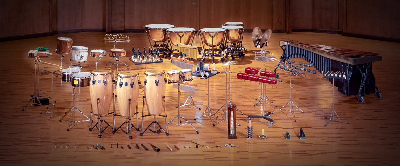 Full percussion orchestra set on a wooden stage, featuring timpani, drums, congas, marimba, cymbals, bells, and various percussion instruments.