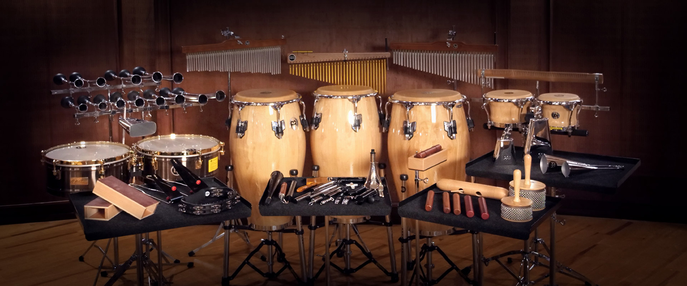 Orchestral percussion setup featuring drum set, congas, bongos, chimes, tambourines, wood blocks, and various shakers and rhythm instruments.