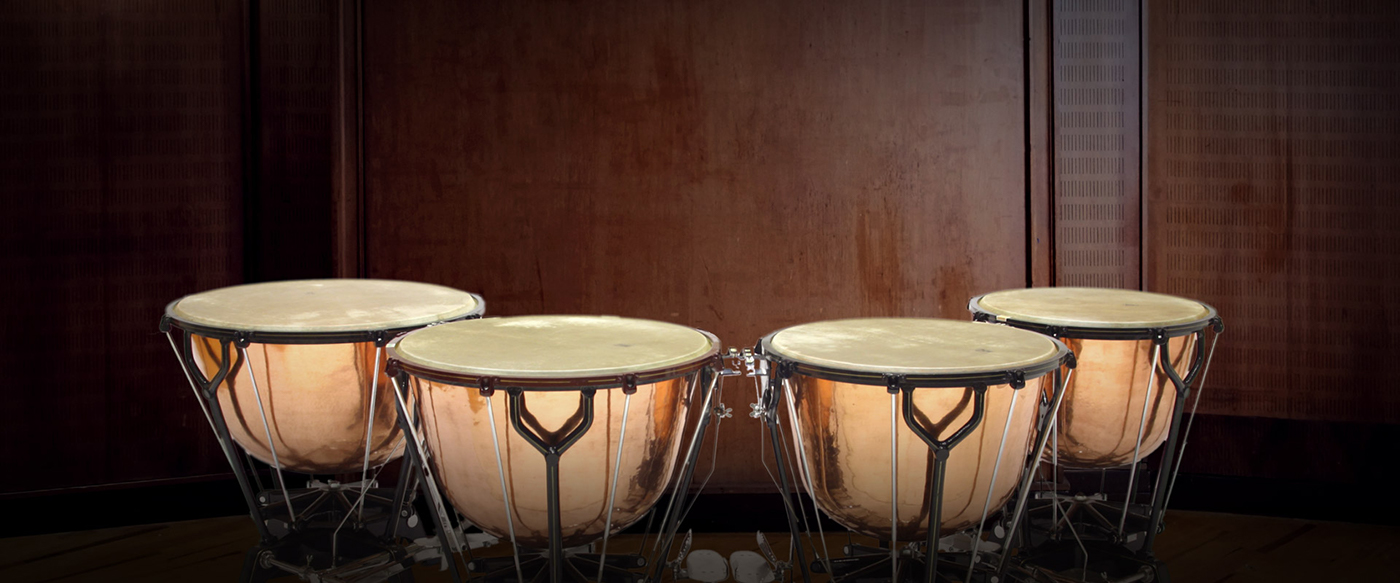 Four concert timpani drums in varying sizes with copper bowls and white drumheads, set against a dark wooden stage background.