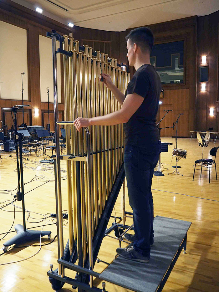 Musician playing a large set of golden tubular bells (chimes) with mallets in a spacious recording studio with wood paneling and musical equipment.