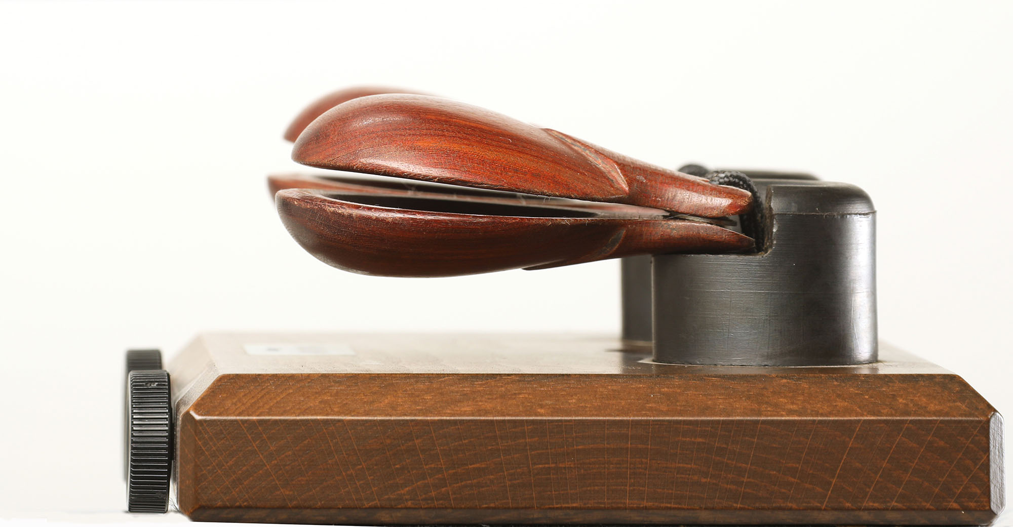 Close-up view of a dark wood castanet machine or holder, featuring two reddish-brown castanets held in playing position on a wooden base with a black dial and dark posts