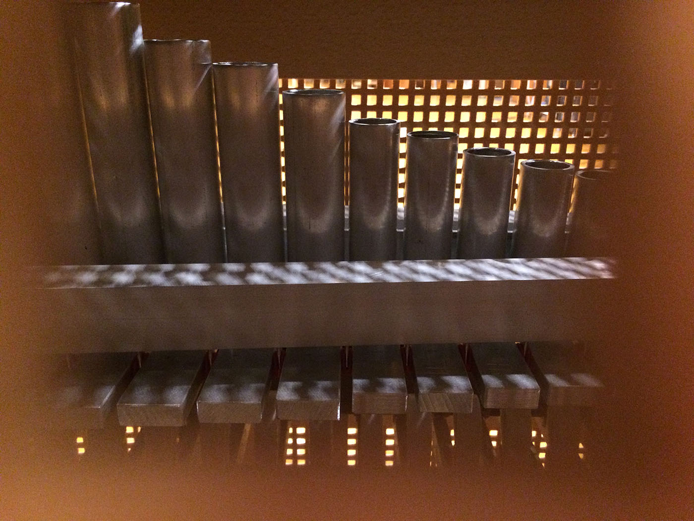 Close-up view of pipe organ interior showing metal pipes of varying sizes, illuminated by a grid pattern of light.