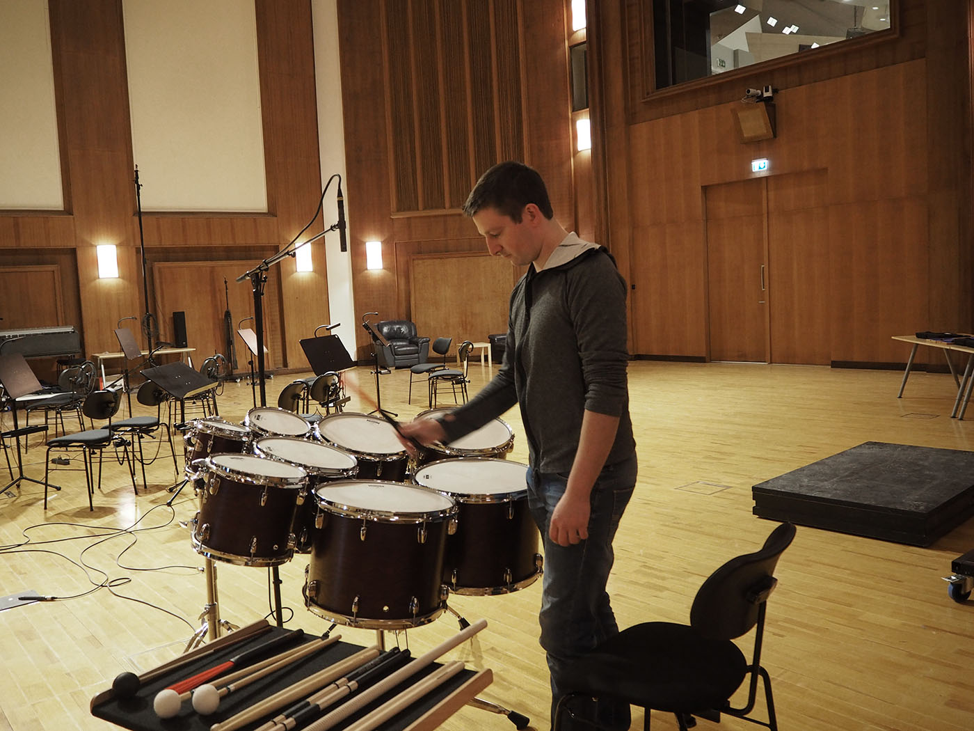 Percussionist striking drums in a large music practice room or recording studio with wooden walls and flooring, sound equipment, and chairs.