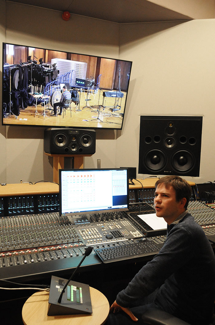 A male audio engineer sits at a sound mixing board in a professional recording studio, looking at a large monitor displaying live studio footage of a musician playing percussion instruments.