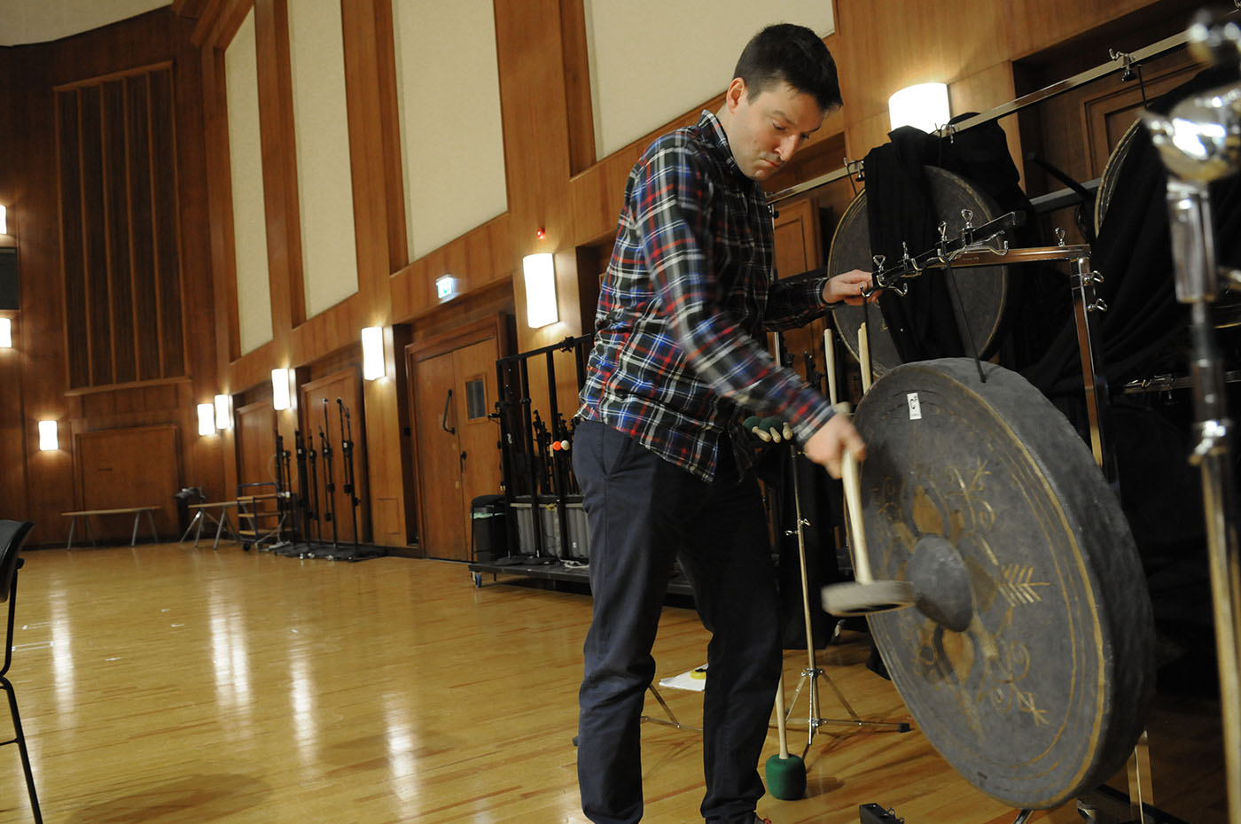 Man in plaid shirt plays a large gong with a mallet in a spacious recording studio with wooden walls and floors.