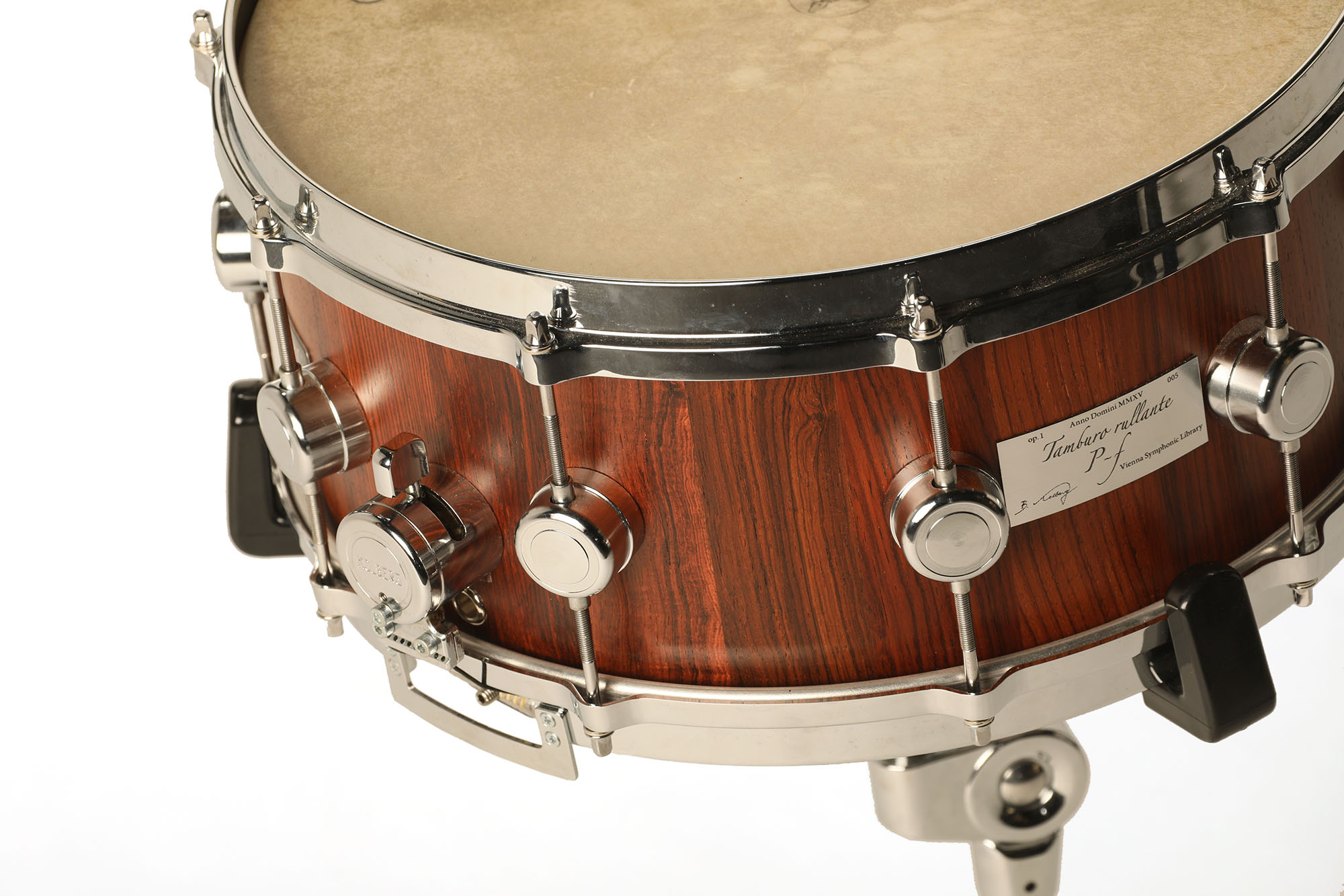 Close-up of a Tamburo Rullante snare drum with rich brown wood grain, chrome hardware, and a light-colored drumhead. A label on the drum reads Anno Domini MMXV Tamburo Rullante, P-F Vienna Symphonic Library.
