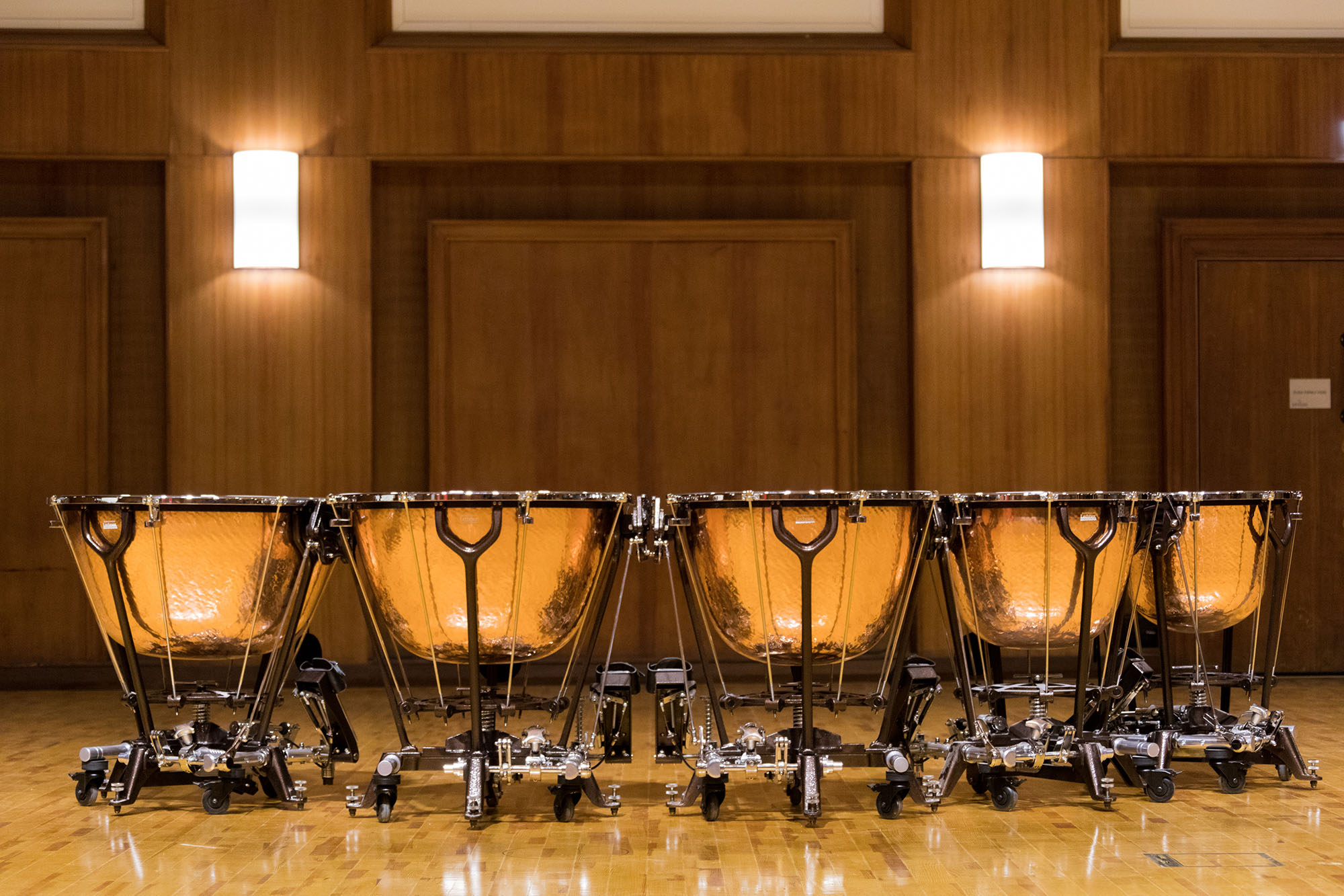 Five copper timpani drums arranged in a row on a wooden stage, with wooden paneling and two illuminated sconces in the background.