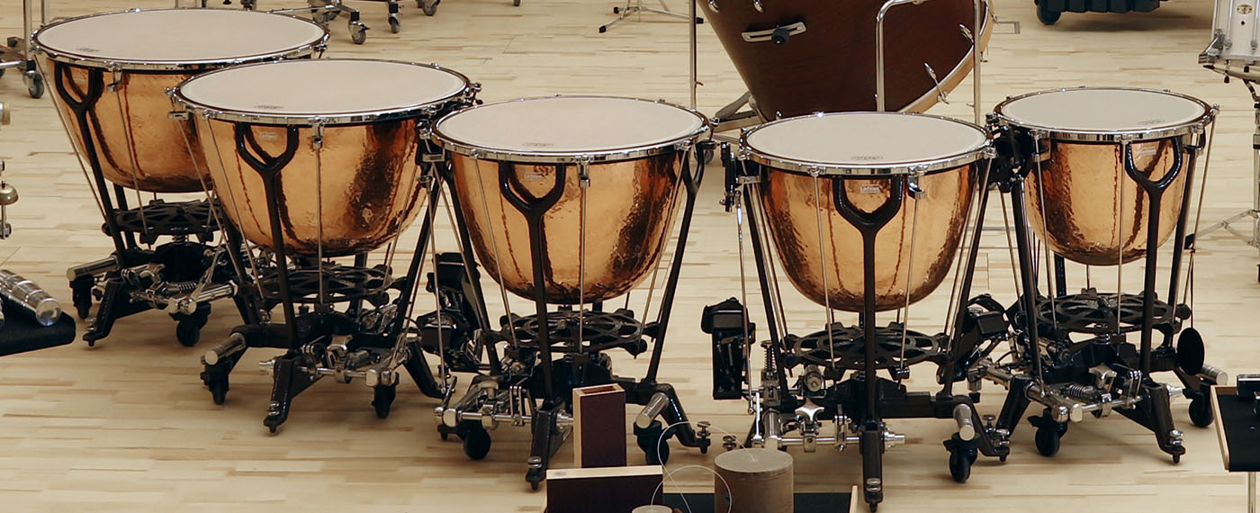 Five copper timpani, also known as kettle drums, arranged in a semicircle on a light wooden floor, ready for an orchestra or band performance.