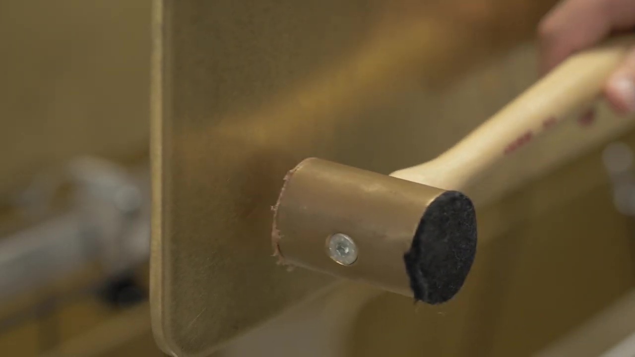 Close-up of a rubber mallet with a wooden handle hitting a golden metal sheet, possibly for fabrication or craftsmanship.