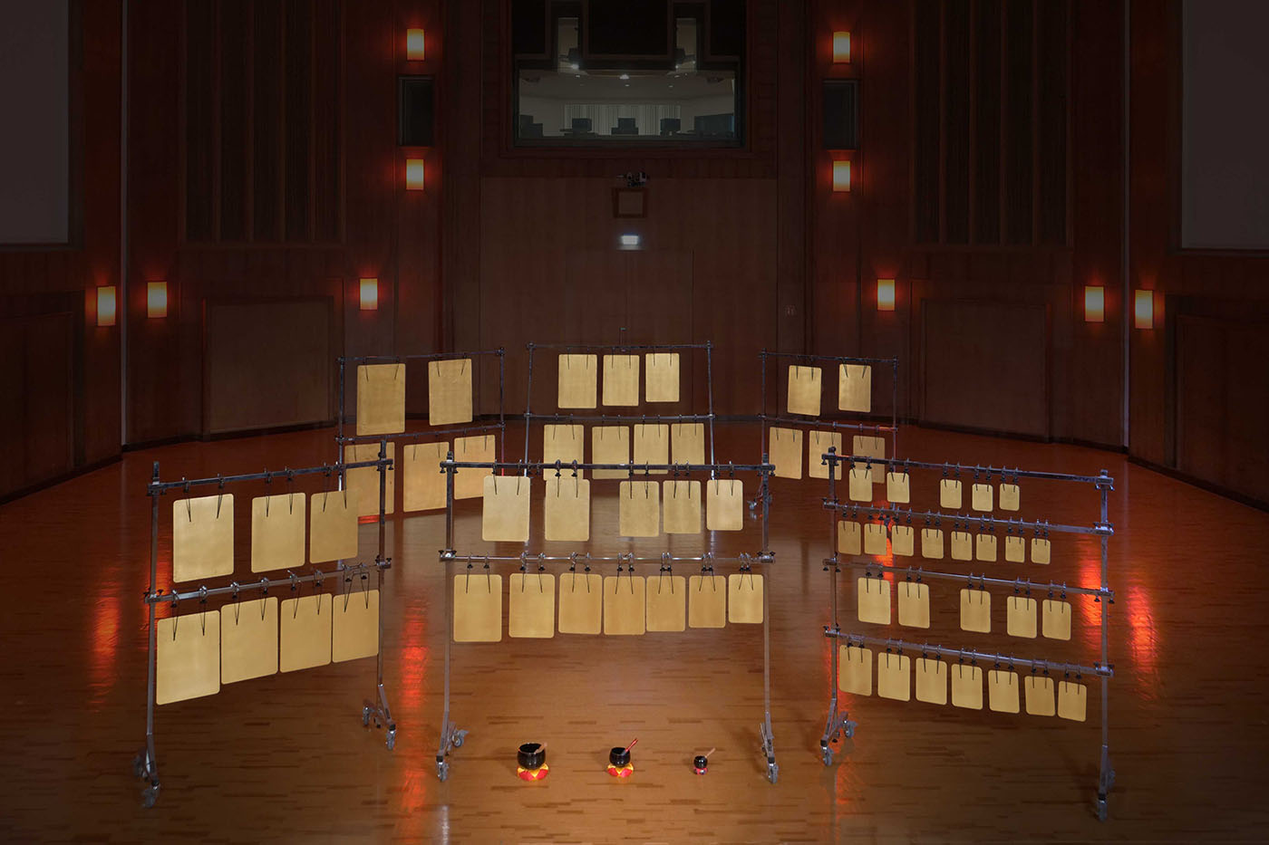 Metallic percussion instruments, possibly Japanese Gongs or bronze plates, on wheeled stands in a dimly lit concert hall or studio with warm wall sconces, ideal for sound recording.