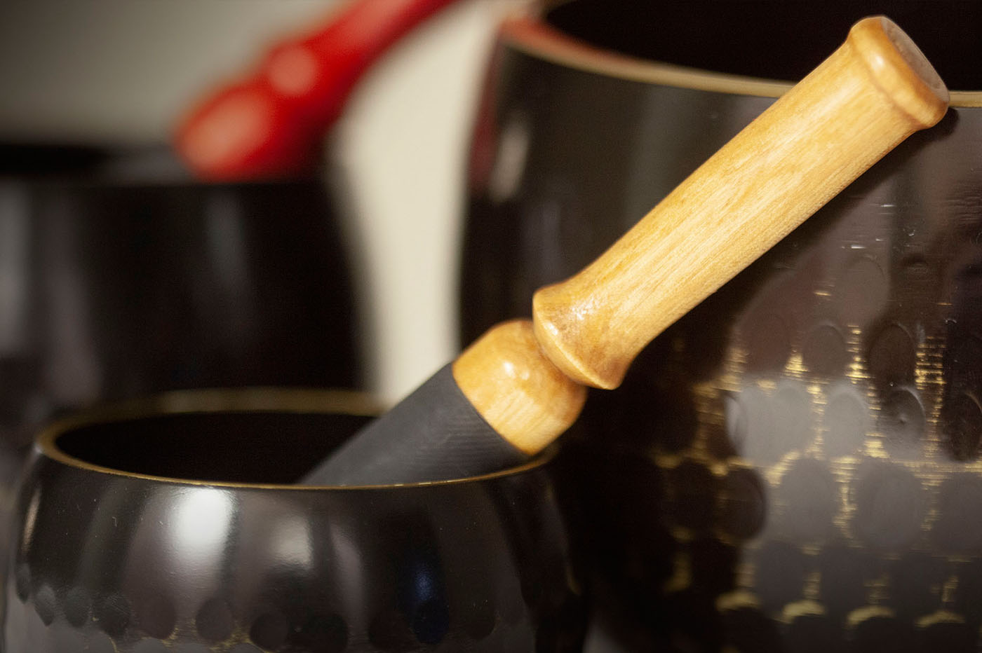 Close-up of a wooden mallet resting inside a black and gold hammered metal singing bowl, with another bowl visible in the background.