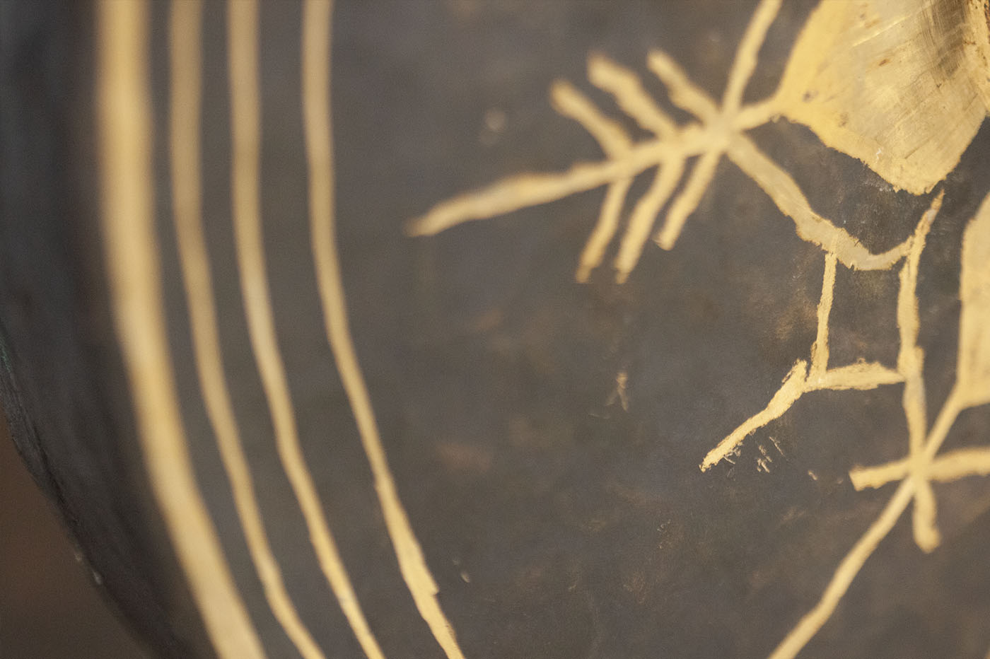 Close-up of a dark brown or black surface with golden-yellow patterns. The patterns consist of vertical lines on the left and abstract, possibly ancient, symbols resembling an arrowhead or a stylized animal bone structure on the right.