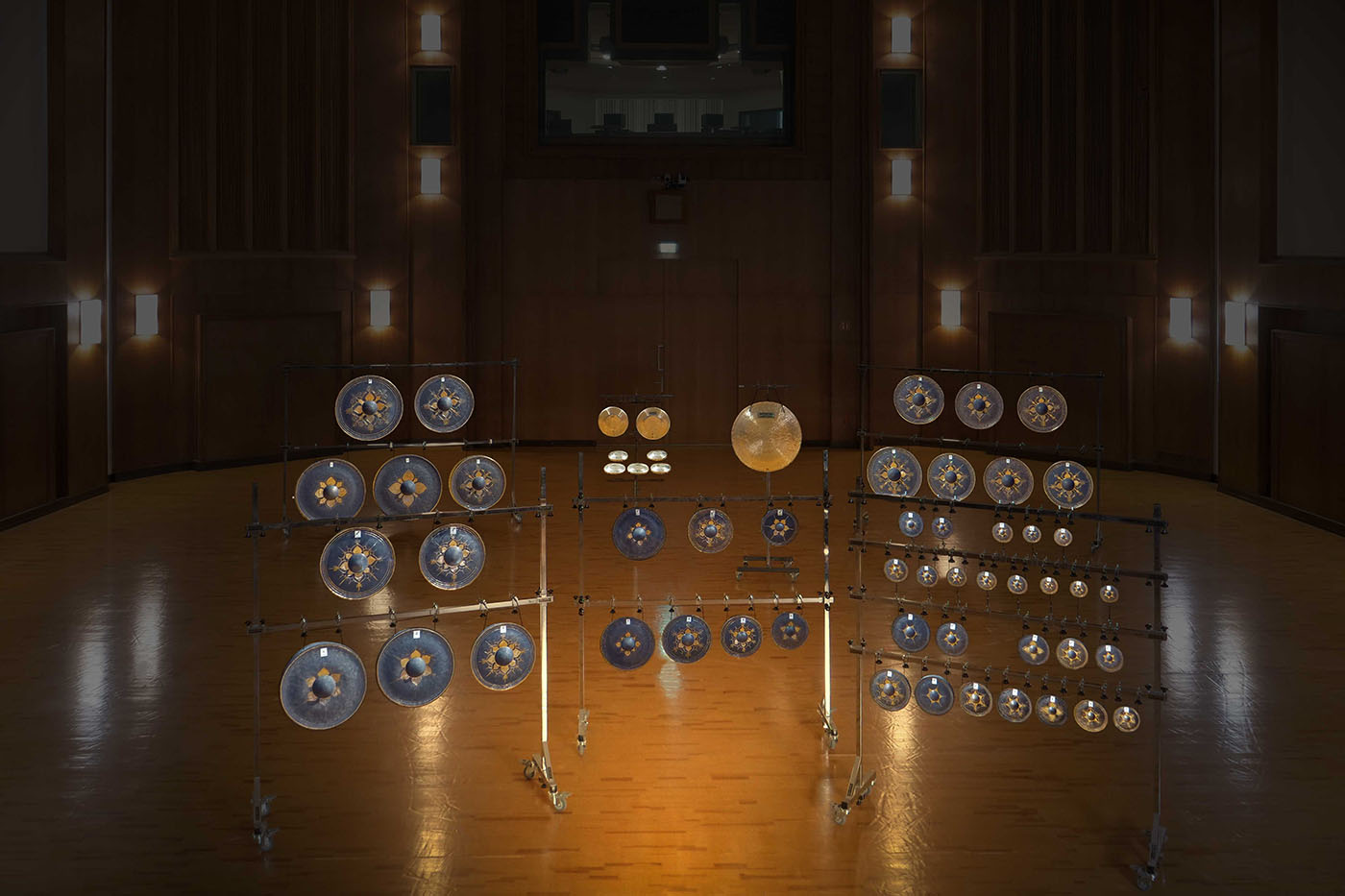 Orchestral gongs and cymbals on stands in a professional concert hall setting, illuminated by warm stage lighting.