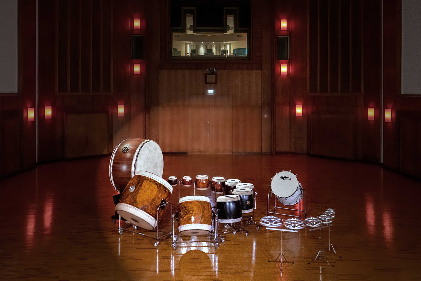 Traditional Japanese taiko drums and percussion instruments arranged on a polished wooden stage in a warm, illuminated hall, with a control booth visible upstairs.