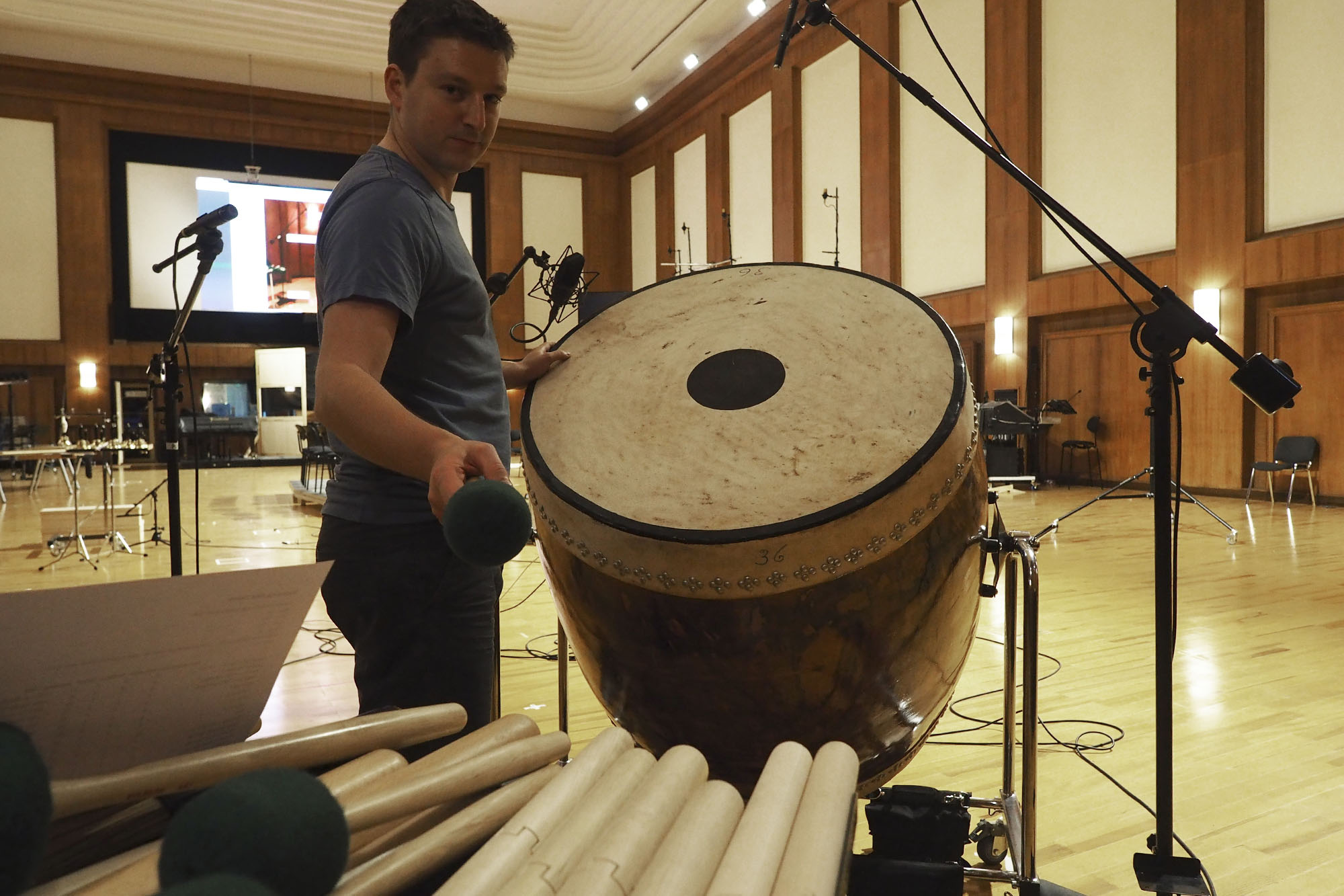 Musician in a recording studio holding a mallet, ready to play a large drum (gong drum) surrounded by percussion instruments and microphones.
