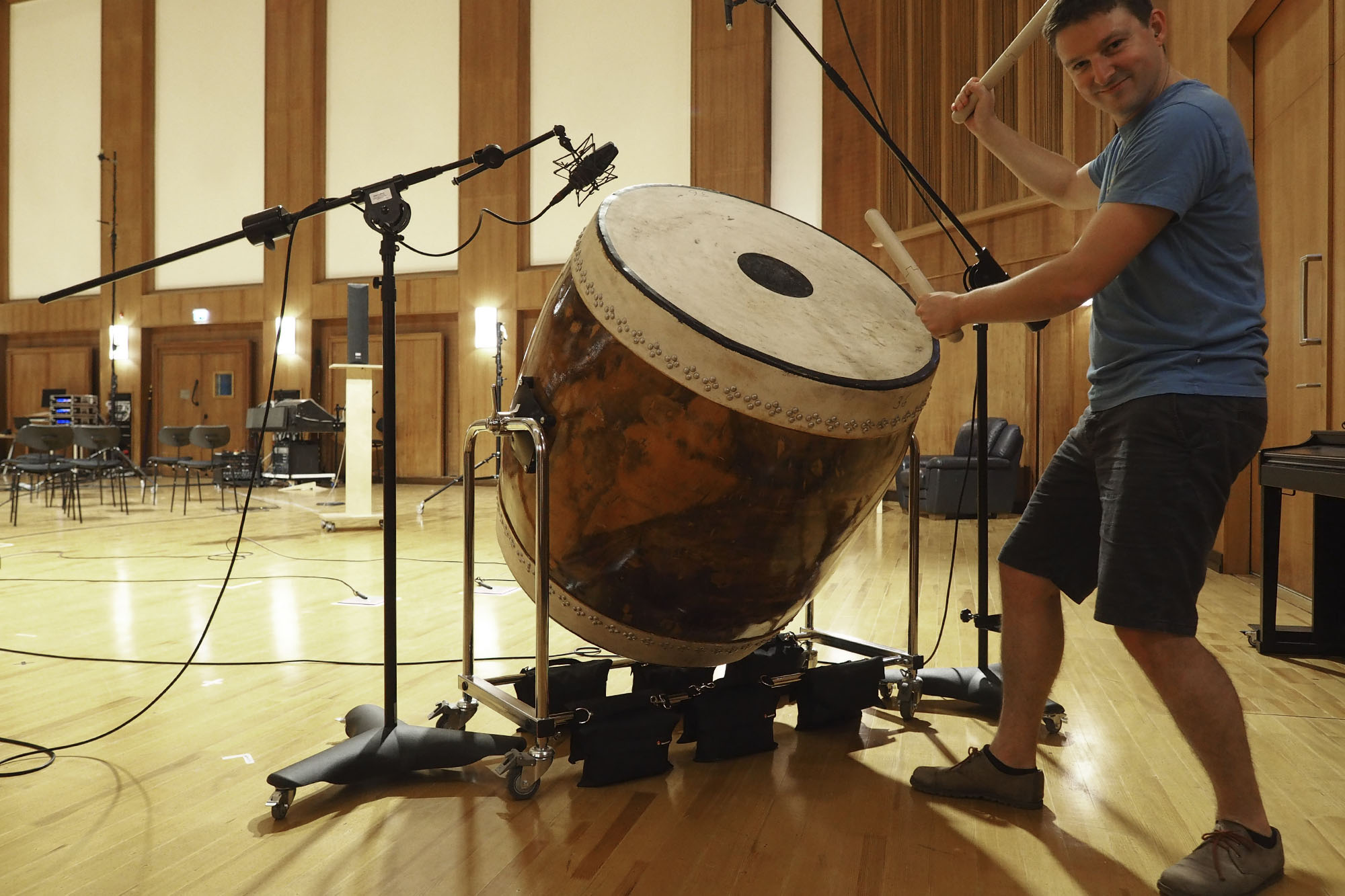Man playing large frame drum in recording studio, with microphones set up.