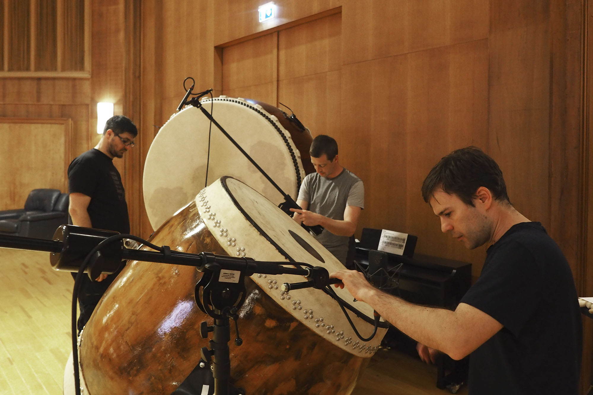 Three sound engineers record immense taiko drums in a professional recording studio with wooden walls