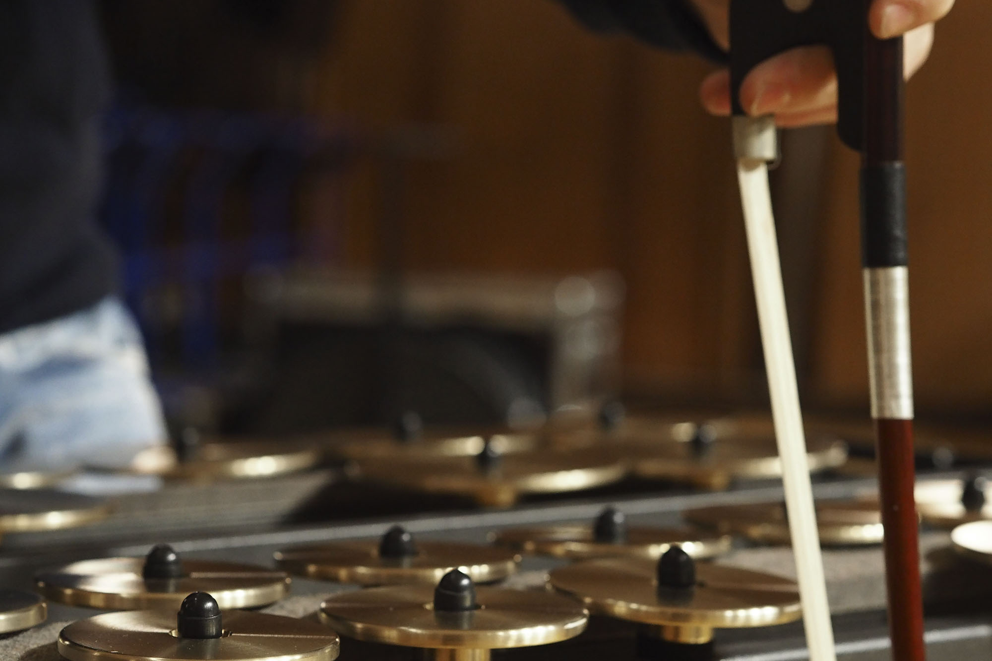 Close-up of a hand holding a bow over a row of brass or gold-colored musical instruments, possibly tuned percussion or gongs, with black centers.