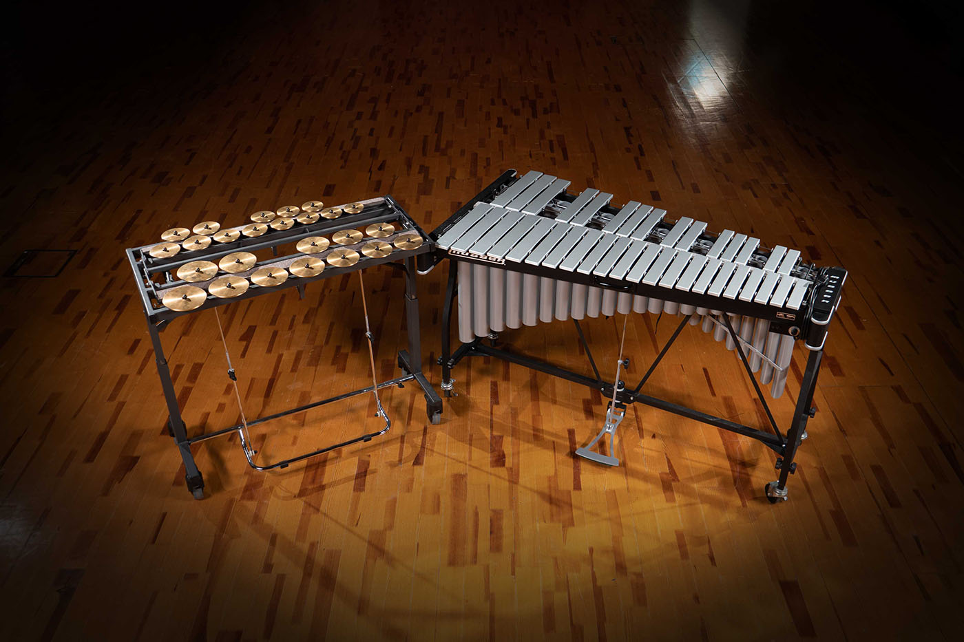 Two sophisticated percussion instruments, a concert vibraphone with silver bars and resonators and a crotale stand with brass cymbals, rest on a polished wooden floor, ready for a musical performance.