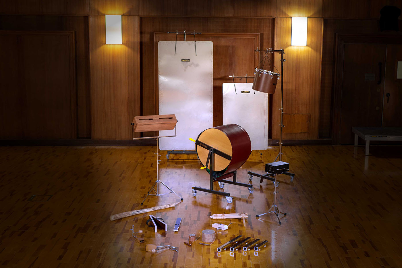 Experimental percussion instruments and sound objects arranged on a wooden floor in a studio setting with acoustic panels.