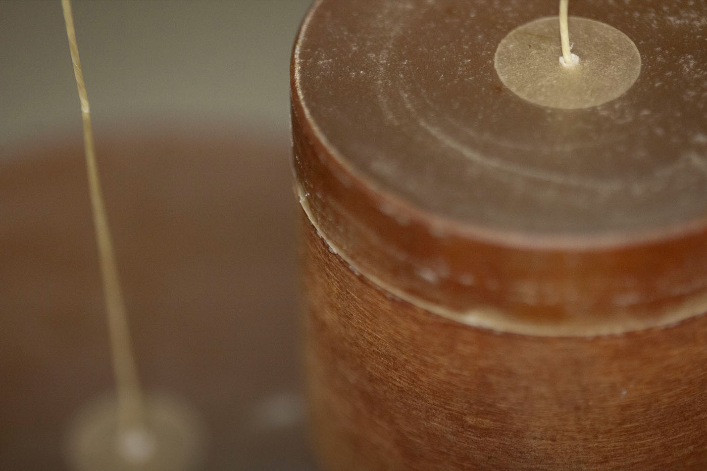 Close-up of a brown candle with a white wick emerging from its top center, surrounded by a light circular wax ring. A thin white thread or strand extends from the bottom of the image upwards, disappearing off-frame.