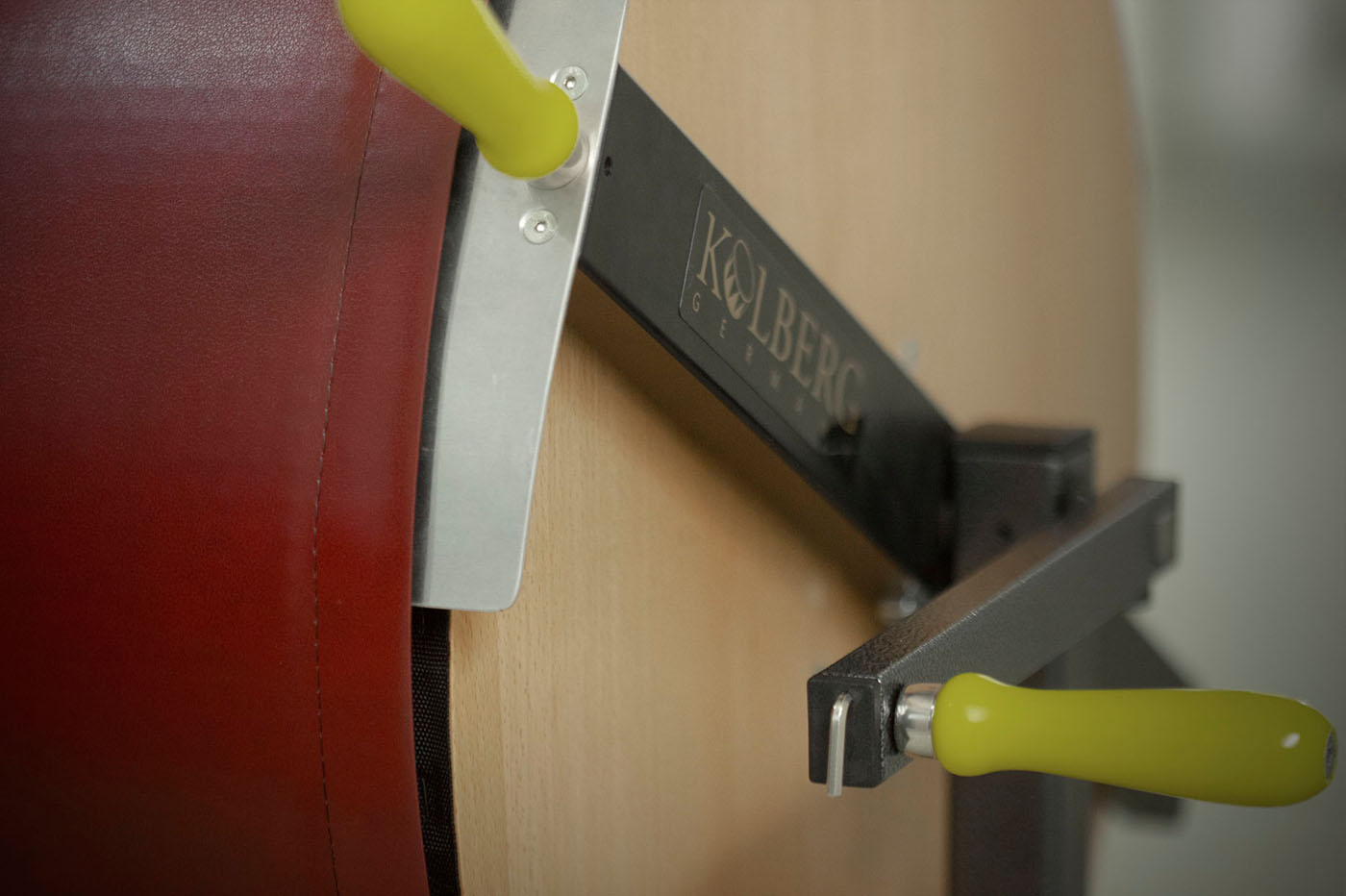 Close-up of a Kolberg Germany percussion instrument or large drum, showing a dark red padded surface, a metallic silver adjustment bracket, a black metal frame with the Kolberg Germany logo, and two bright yellow handles.