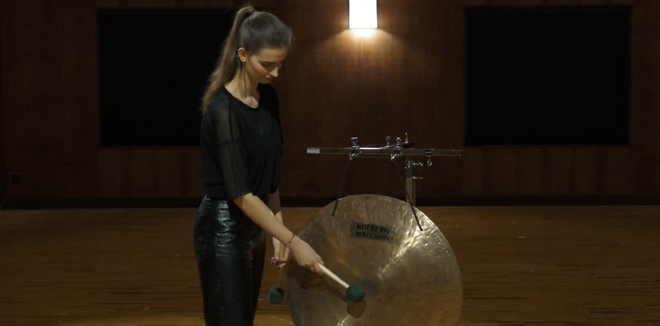 Woman playing a Kolberg Percussion Tam-tam Flat 70, striking the large flat gong with mallets in a dimly lit room representing musical performance.