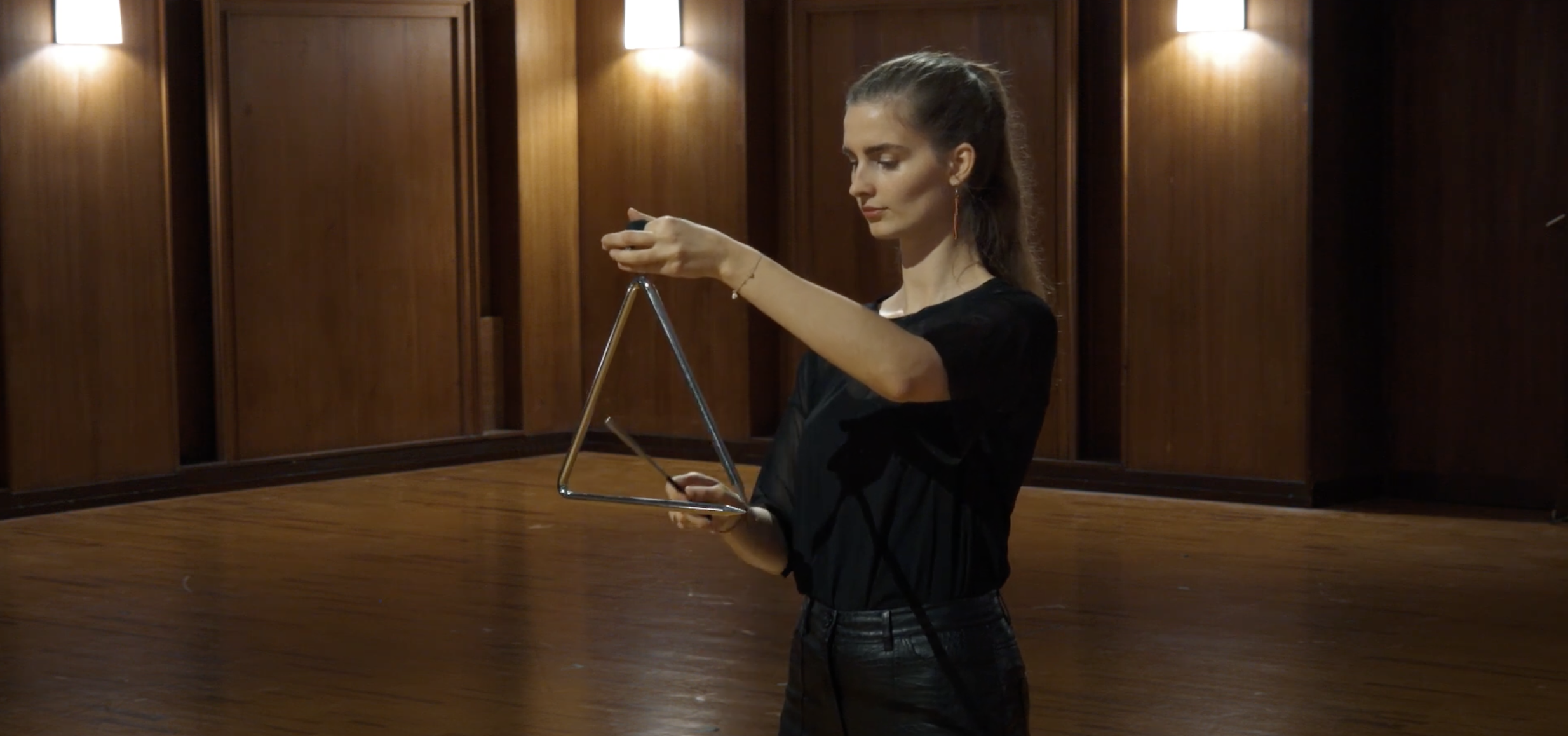 Woman playing a 30 cm steel triangle instrument in a room with wood paneling.