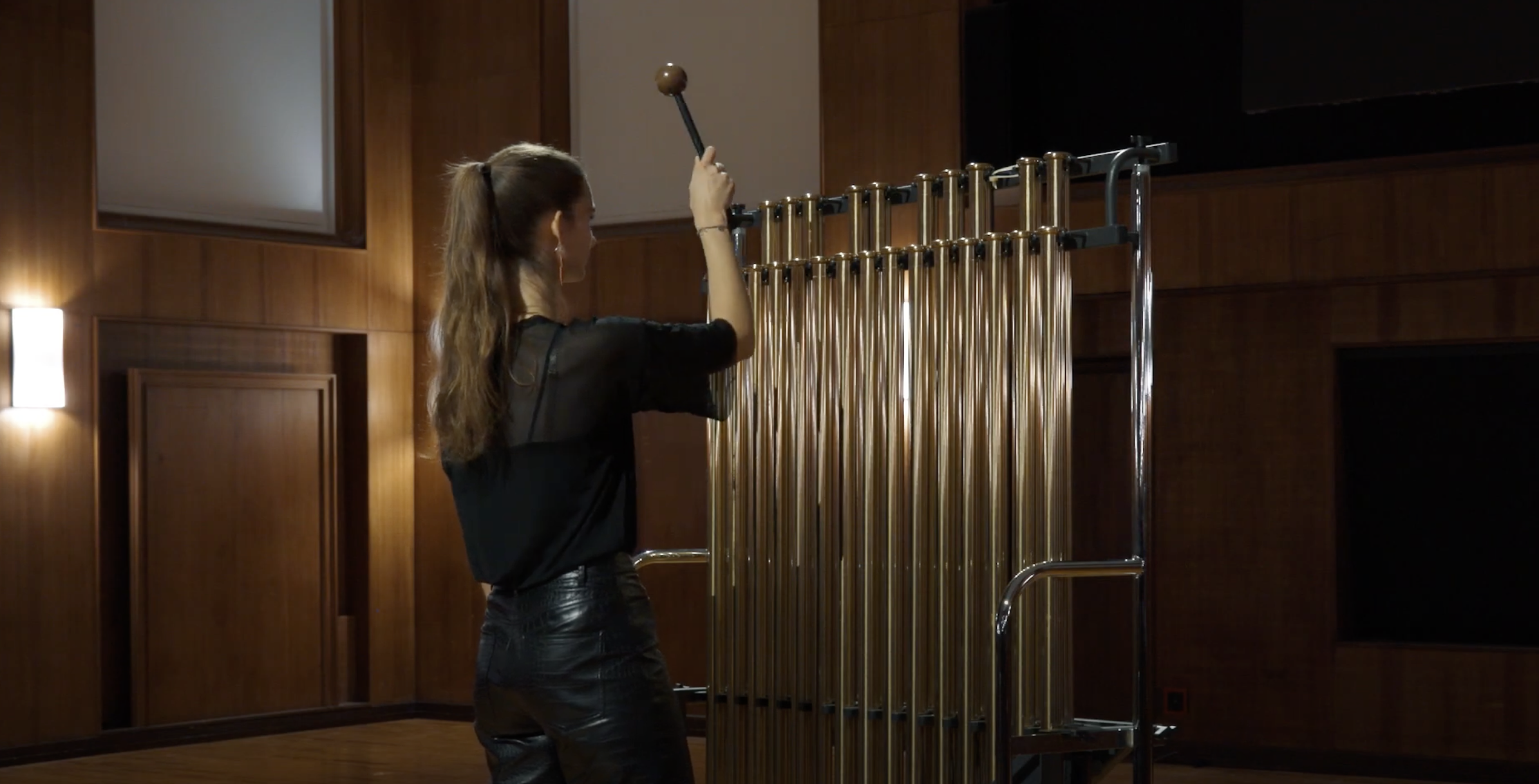 Musician playing Bergerault C25 F4-F6 tubular bells, a large percussion instrument with tall brass tubes, in a wood-paneled room.