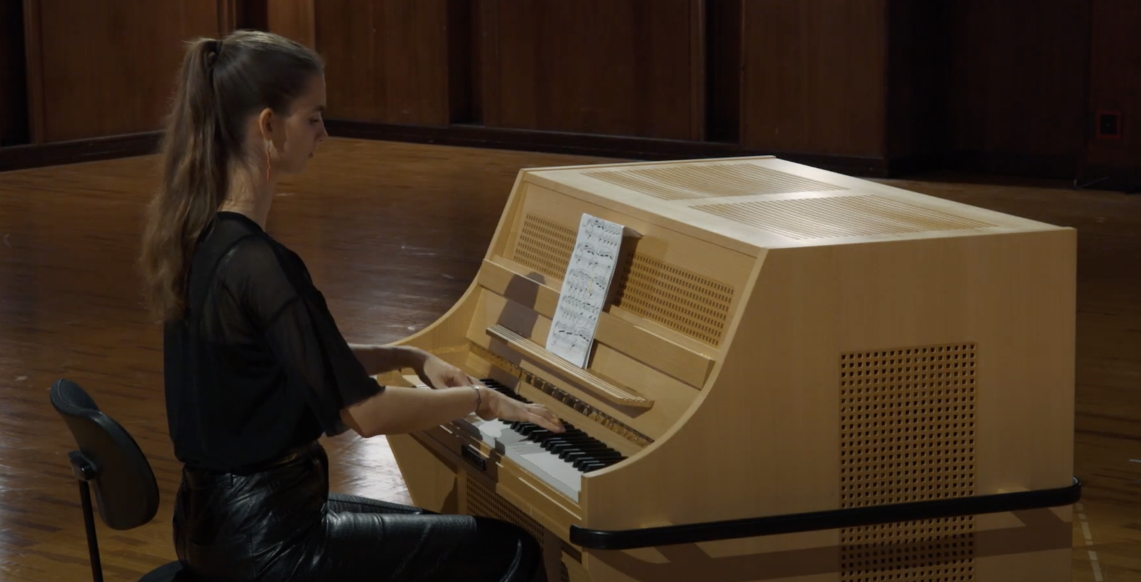 Young woman, Celesta Kolberg, playing a natural wood tone celesta with sheet music on a stand, performing a musical piece.