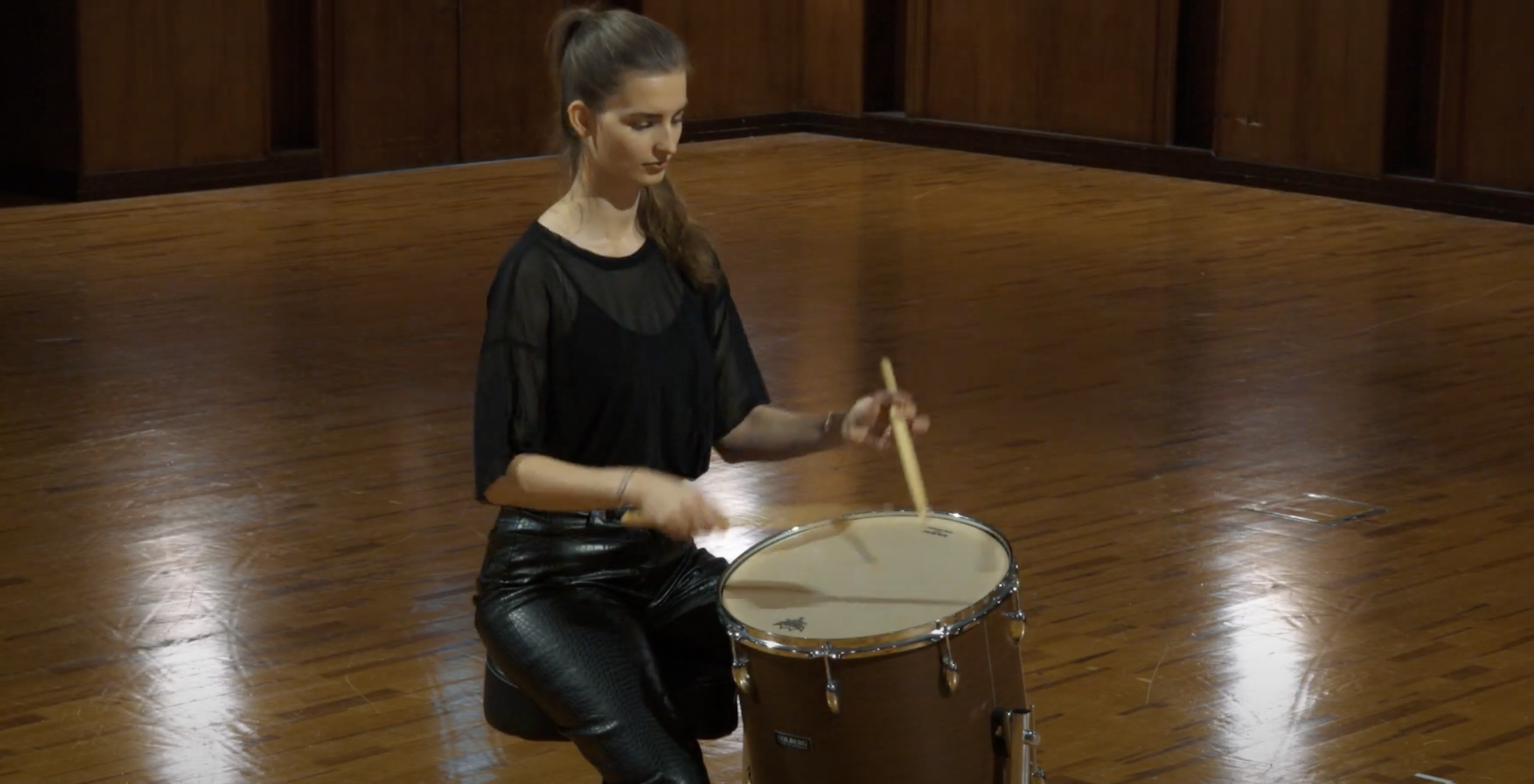 Young woman playing a Kolberg 16x16 Field Drum with drumsticks in a hall with wooden floors.