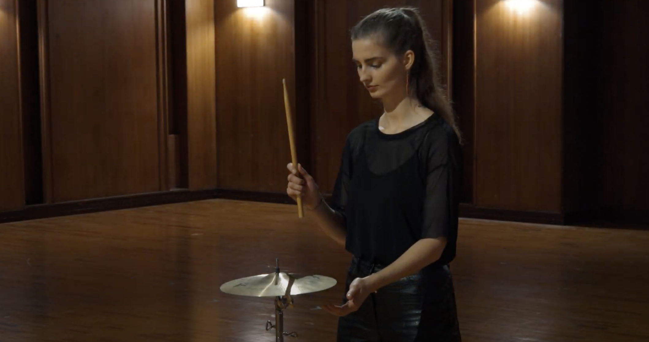 Young woman in black playing Zildjian Avedis 12-inch cymbal with drumstick in a wooden room.