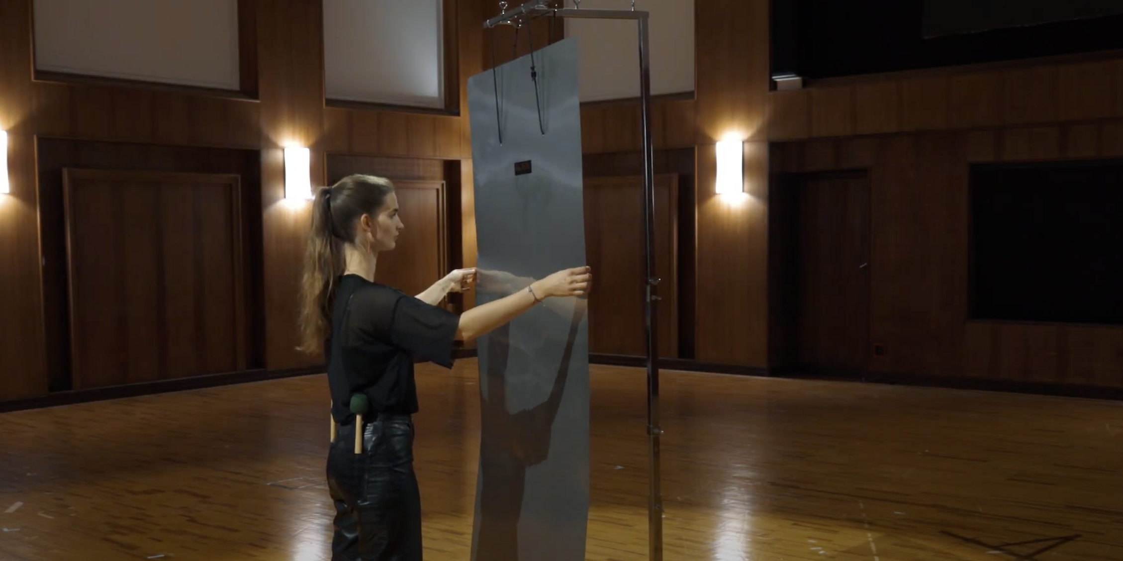 Young woman holding a large thundersheet, demonstrating the percussion instrument in a wood-paneled room.